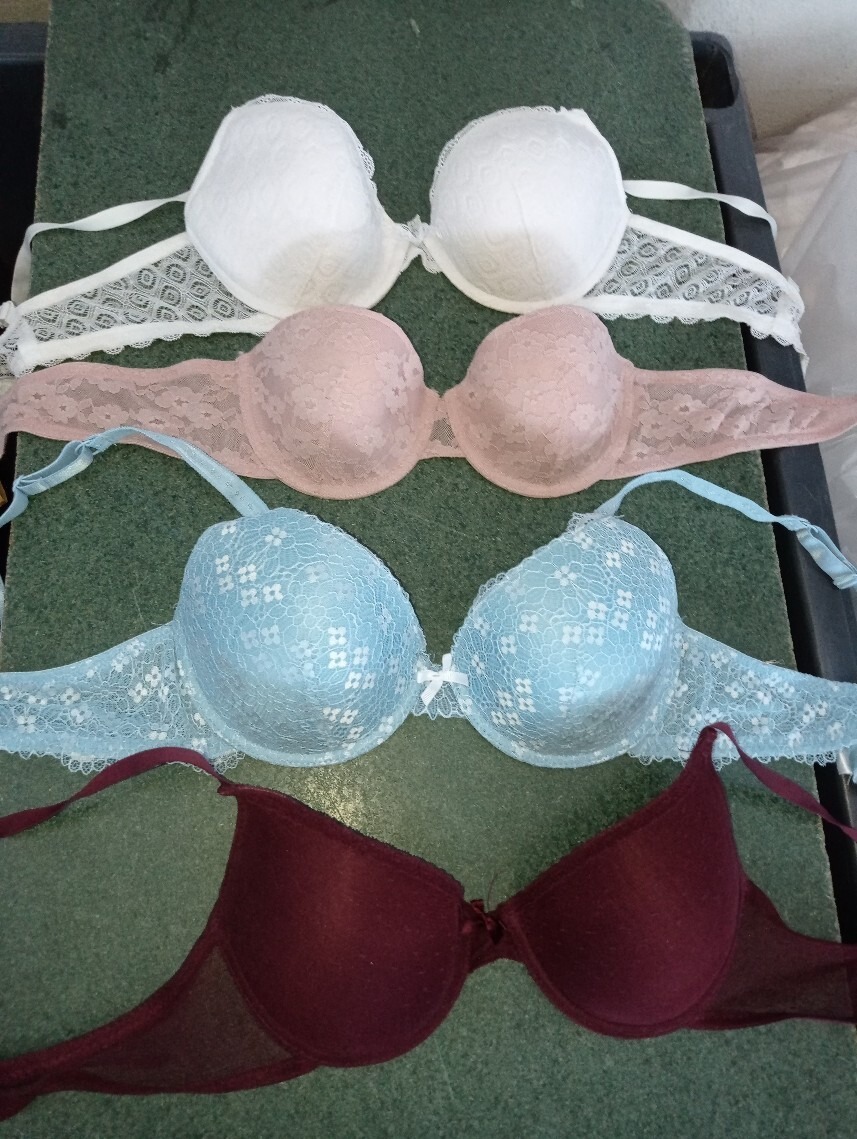 Cztery używane biustonosze w różnych kolorach (biały, różowy, błękitny, bordowy) ułożone jeden na drugim, każdy z koronkowymi detalami, na zielonym tle.
