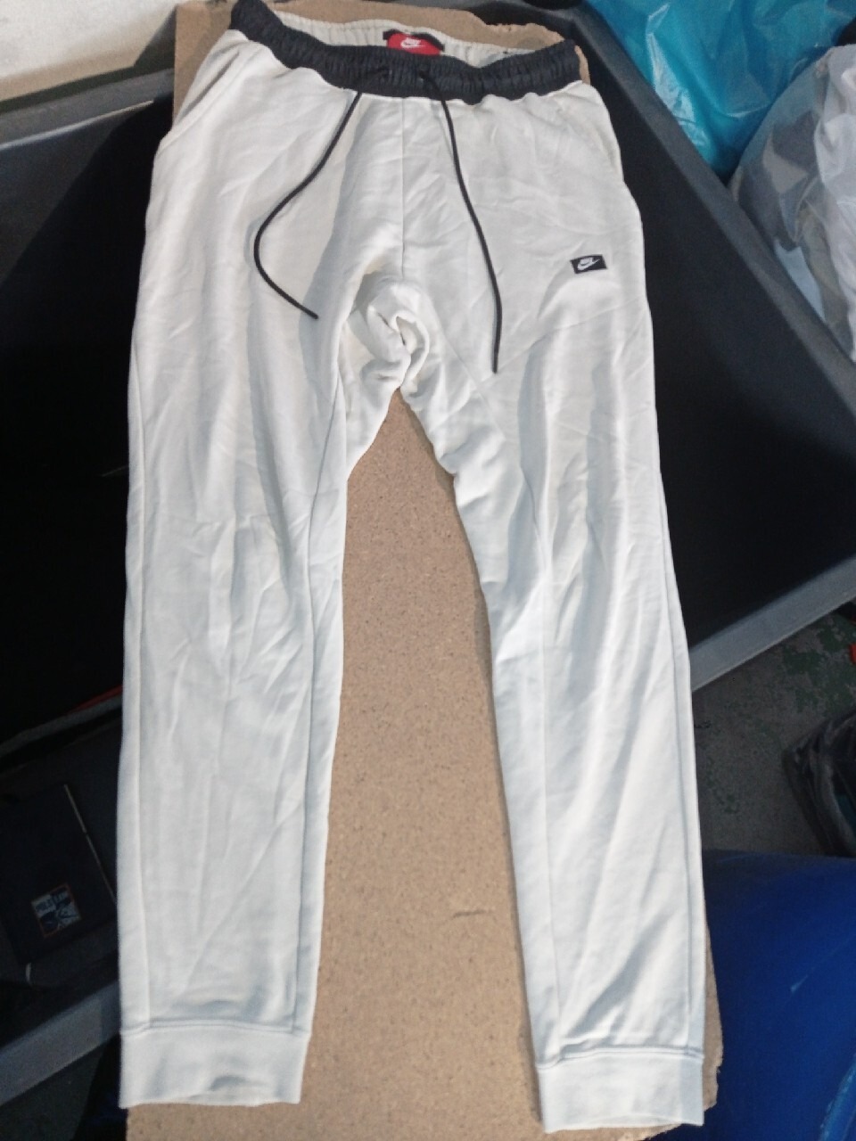 Jasnoszare dresowe spodnie Nike z czarnym ściągaczem w pasie i sznurkiem, leżące na kartonowym podłożu w nieuporządkowanym otoczeniu.