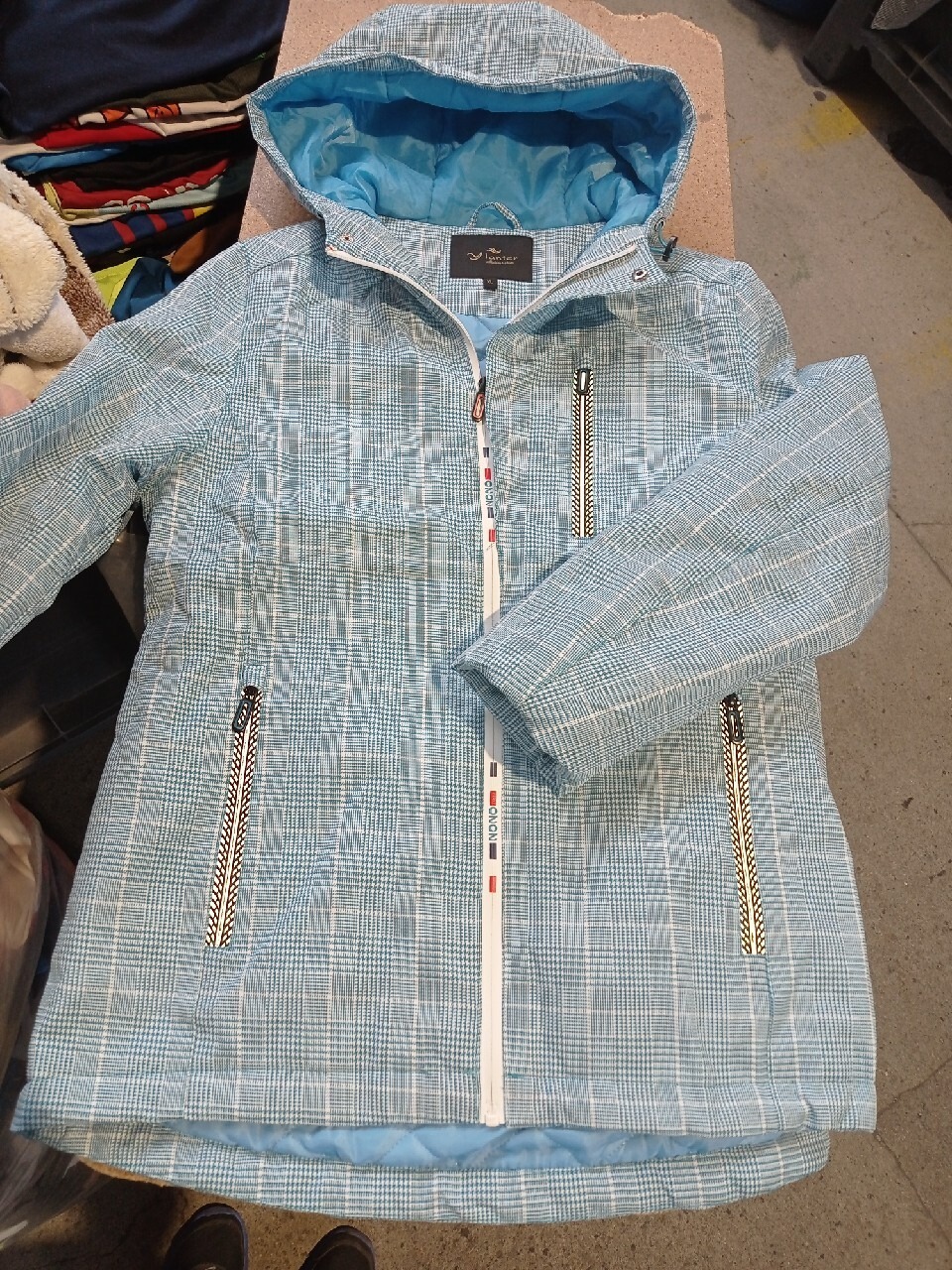 Jasnoniebieska kurtka w drobną kratę z kapturem, zamek błyskawiczny z dekoracyjnymi elementami i podszewką w kolorze błękitnym.