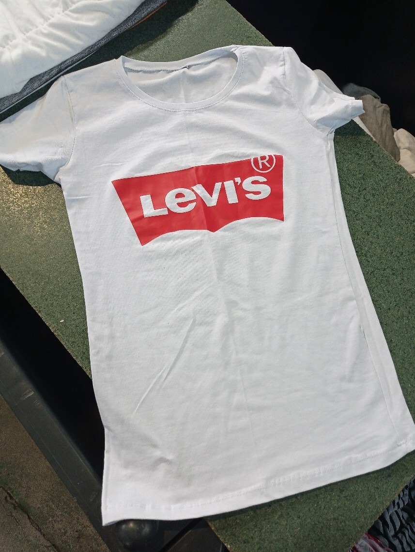 Biała koszulka z krótkim rękawem i dużym czerwonym logo Levi's na przodzie, leżąca na zielonym blacie.