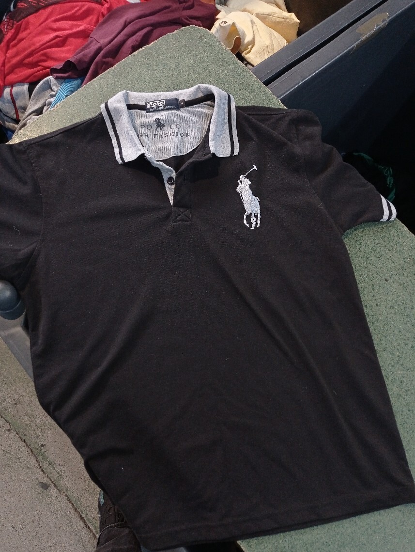 Czarna koszulka polo z szarym kołnierzykiem i białym logo jeźdźca Polo Ralph Lauren, leżąca na szorstkiej powierzchni, w tle widoczne inne ubrania.