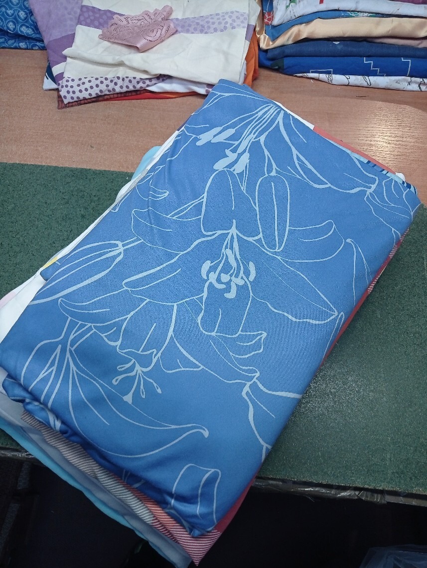 Stos złożonej pościeli z niebieską narzutą w kwiatowy wzór, leżący na zielonym blacie, z innymi tekstyliami w tle.