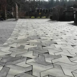 Ułożona nawierzchnia z szarej kostki o unikalnym wzorze, z widocznymi drobnymi kamykami w szczelinach, prowadząca do otwartej, metalowej bramy wjazdowej i domu w tle.