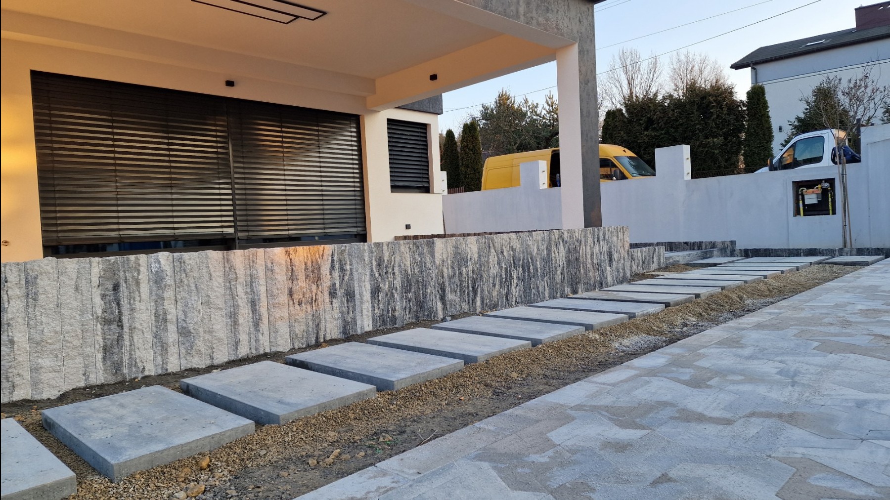 Nowoczesna ścieżka z kwadratowych płyt betonowych prowadząca do domu, z widoczną ścianą z kamienia dekoracyjnego i żwirowym wypełnieniem, na tle białego ogrodzenia i zaparkowanego żółtego vana.