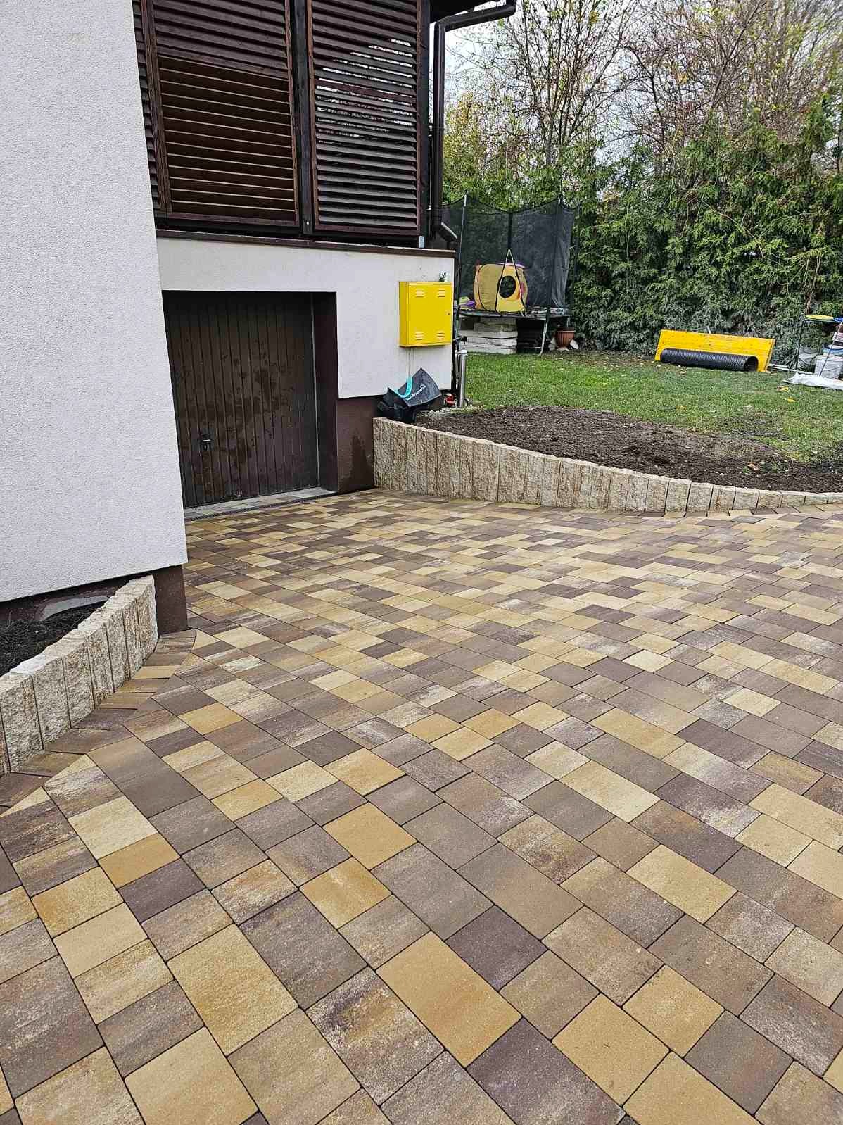 Ułożona kostka brukowa w odcieniach brązu i beżu przed budynkiem z drewnianymi okiennicami i zielonym trawnikiem w tle.