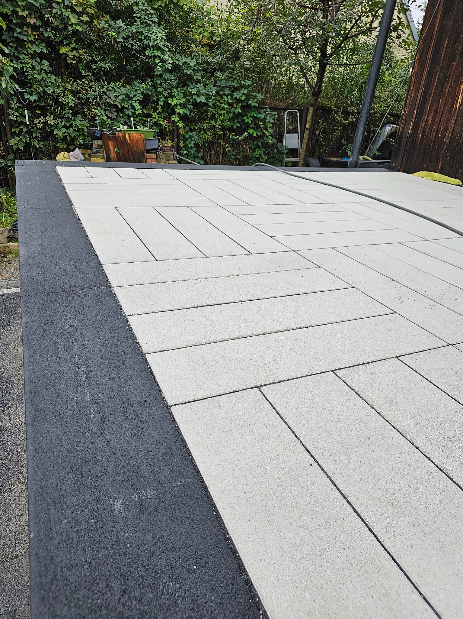 Jasnoszare prostokątne płyty ułożone w jodełkę na nowym chodniku z czarną obwódką, widoczne zieleń i drabina w tle.