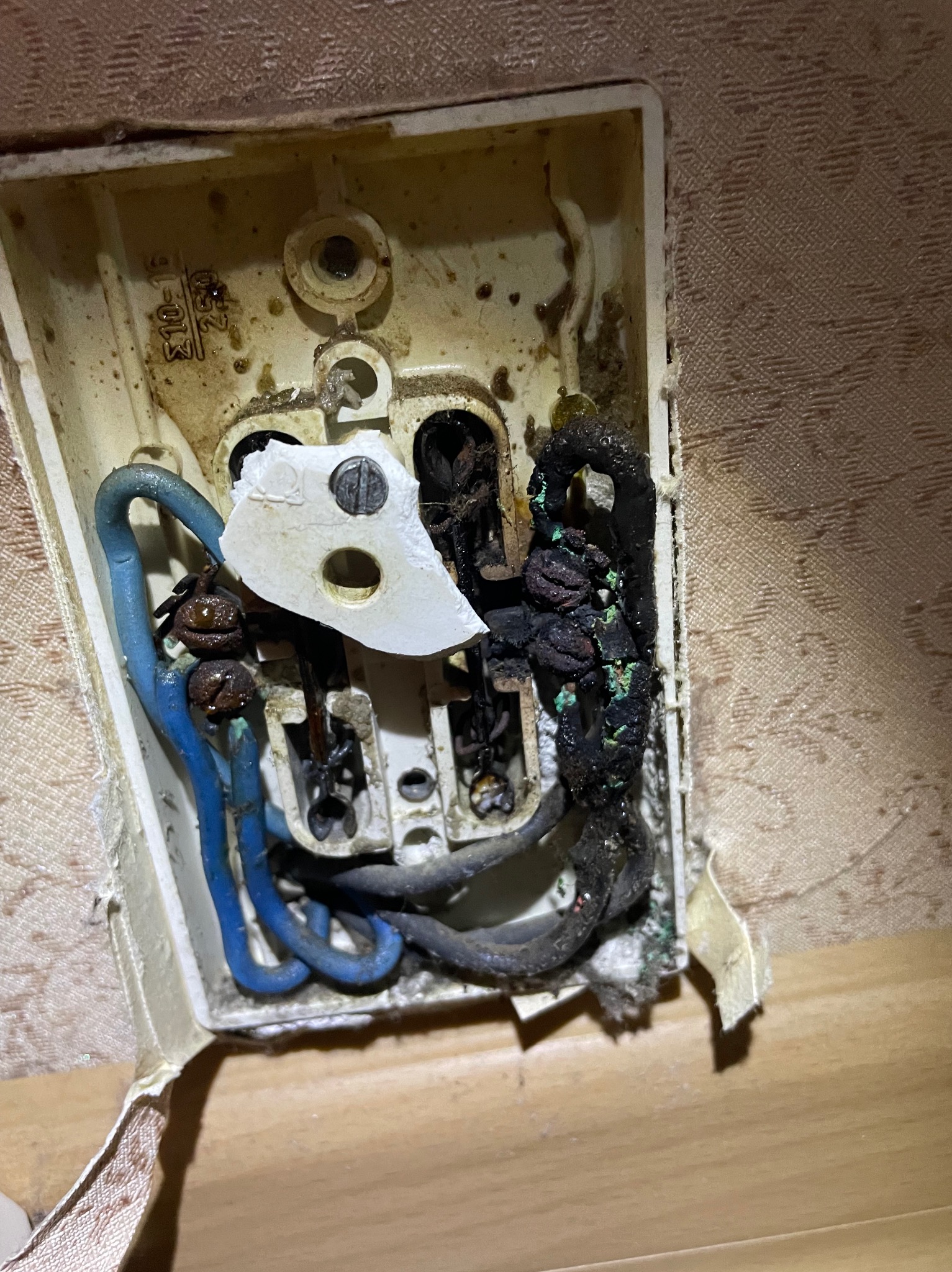 Spalone gniazdo elektryczne z widocznymi uszkodzeniami termicznymi, stopionymi kablami i fragmentem obudowy. Widoczne niebieskie i czarne przewody. Obudowa gniazda jest brudna i częściowo oderwana...