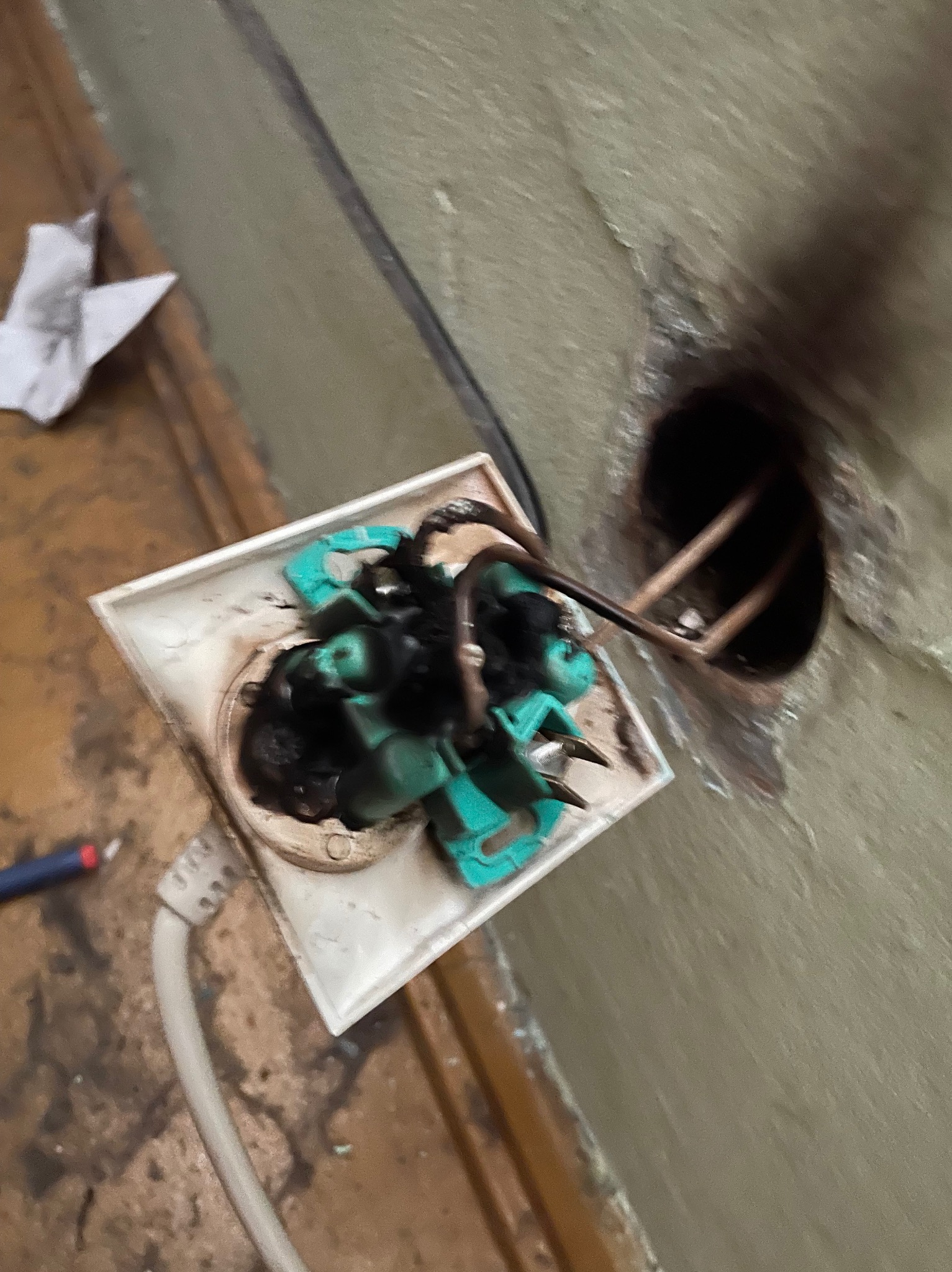 Spalone gniazdo elektryczne z widocznymi stopionymi elementami i okopceniami, wystające przewody, ściana z ubytkiem tynku wokół puszki elektrycznej.