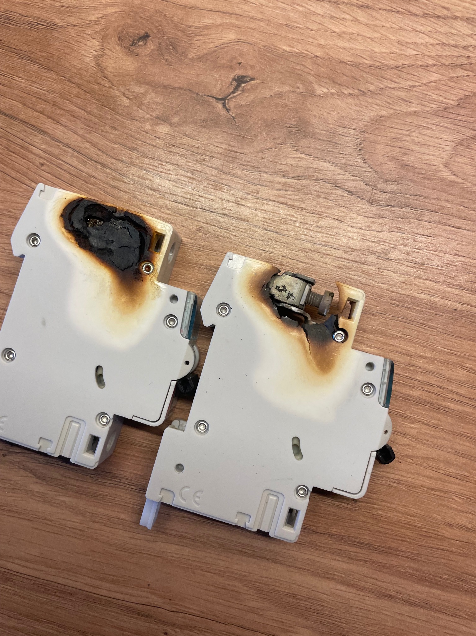 Dwa spalone, białe wyłączniki automatyczne leżące na drewnianej powierzchni, ukazujące stopień uszkodzeń termicznych.