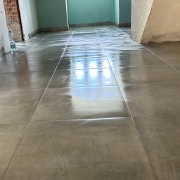 Osoba prywatna - Ułożona, szara, błyszcząca podłoga z płytek w trakcie remontu, z widoczną butelką wody mineralnej przy ścianie i surowymi ścianami w tle.