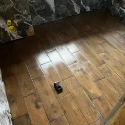 Ułożone płytki podłogowe imitujące drewno w łazience, z widocznym poziomnicą laserową i fragmentem marmurowej ściany z instalacją hydrauliczną.