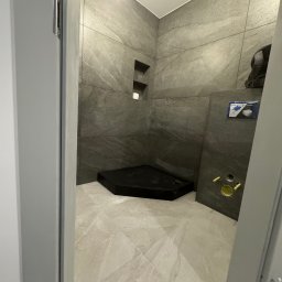 Osoba prywatna - Częściowo wykończona kabina prysznicowa z ciemnoszarą okładziną ścienną imitującą kamień i jasnobeżową podłogą z płytek imitujących marmur, widoczny czarny brodzik i elementy instalacji podtynkowych.