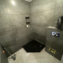 Osoba prywatna - Wnętrze łazienki w trakcie prac wykończeniowych; widoczny czarny brodzik, szare płytki imitujące kamień na ścianach i podłodze, instalacja podtynkowego WC z przyciskiem spłukującym, oraz otwory...