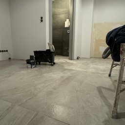 Osoba prywatna - Przygotowania do prac wykończeniowych w Opolu: widok na podłogę z szarych płytek, wiadro z narzędziami malarskimi i drabina w tle łazienki z szarą okładziną.