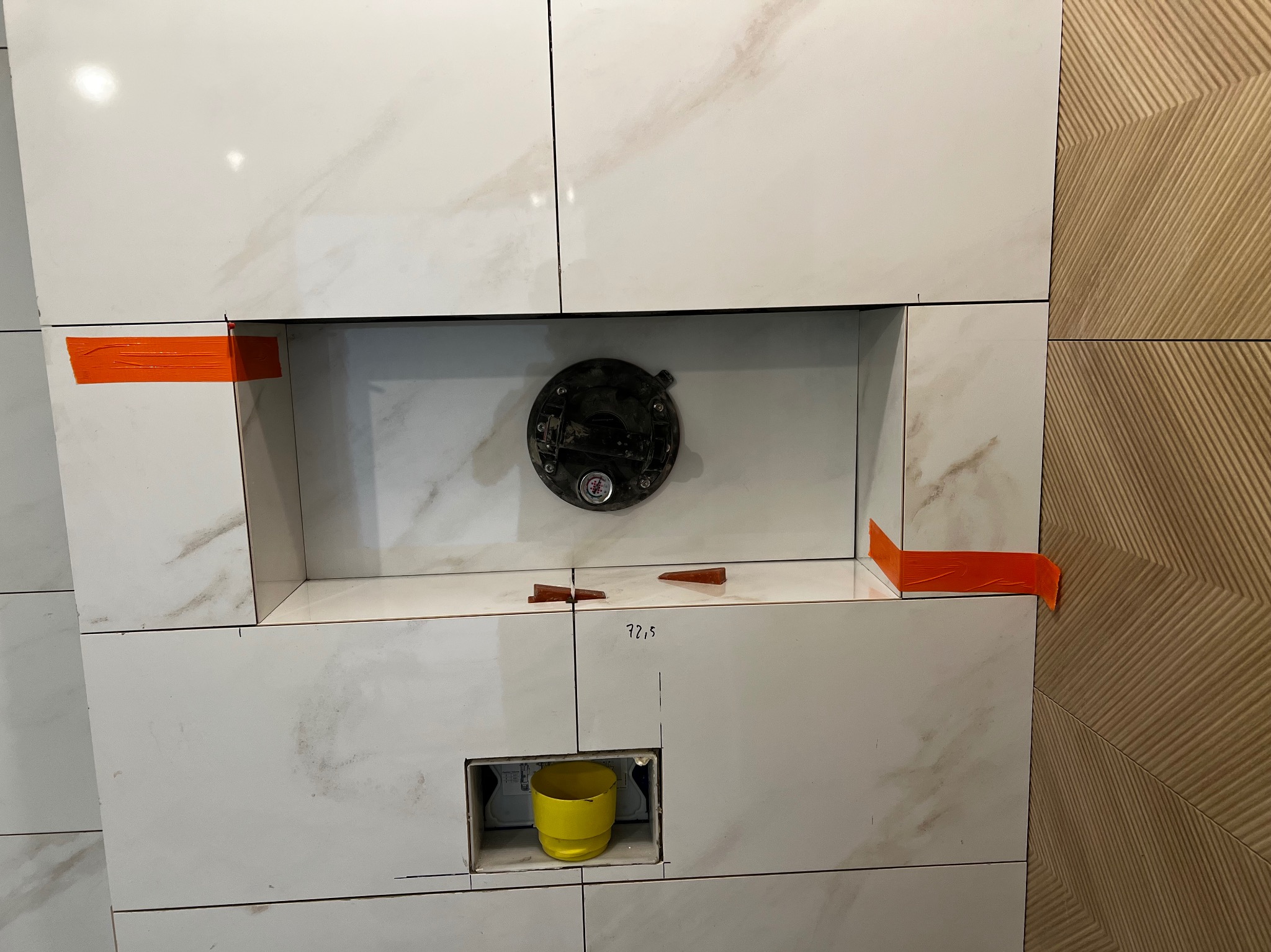 Montaż białych płytek imitujących marmur z pomarańczową taśmą maskującą, klinami poziomującymi i żółtym kubkiem w otworze na instalację podtynkową.
