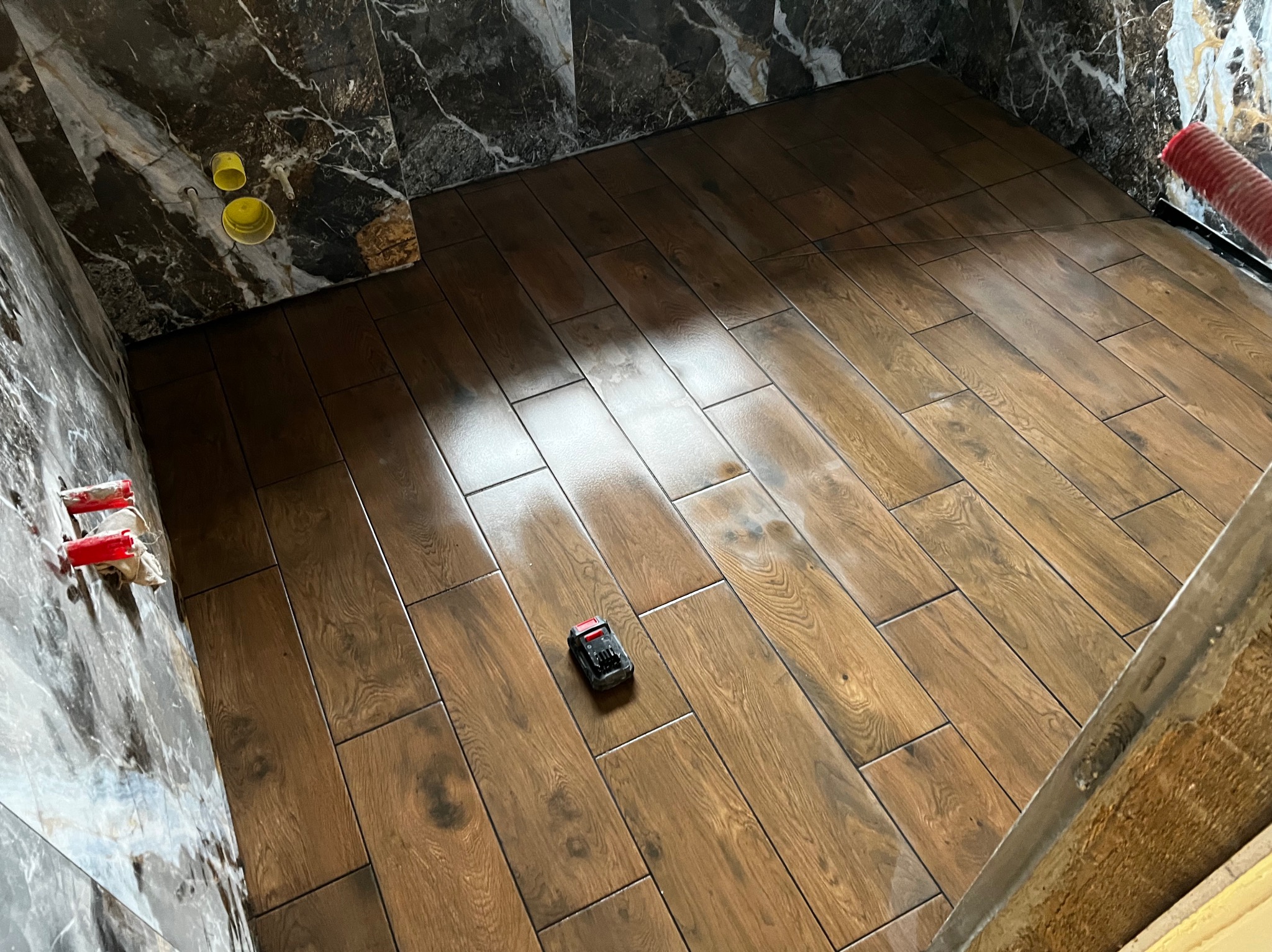 Ułożone płytki podłogowe imitujące drewno w łazience, z widocznym poziomnicą laserową i fragmentem marmurowej ściany z instalacją hydrauliczną.