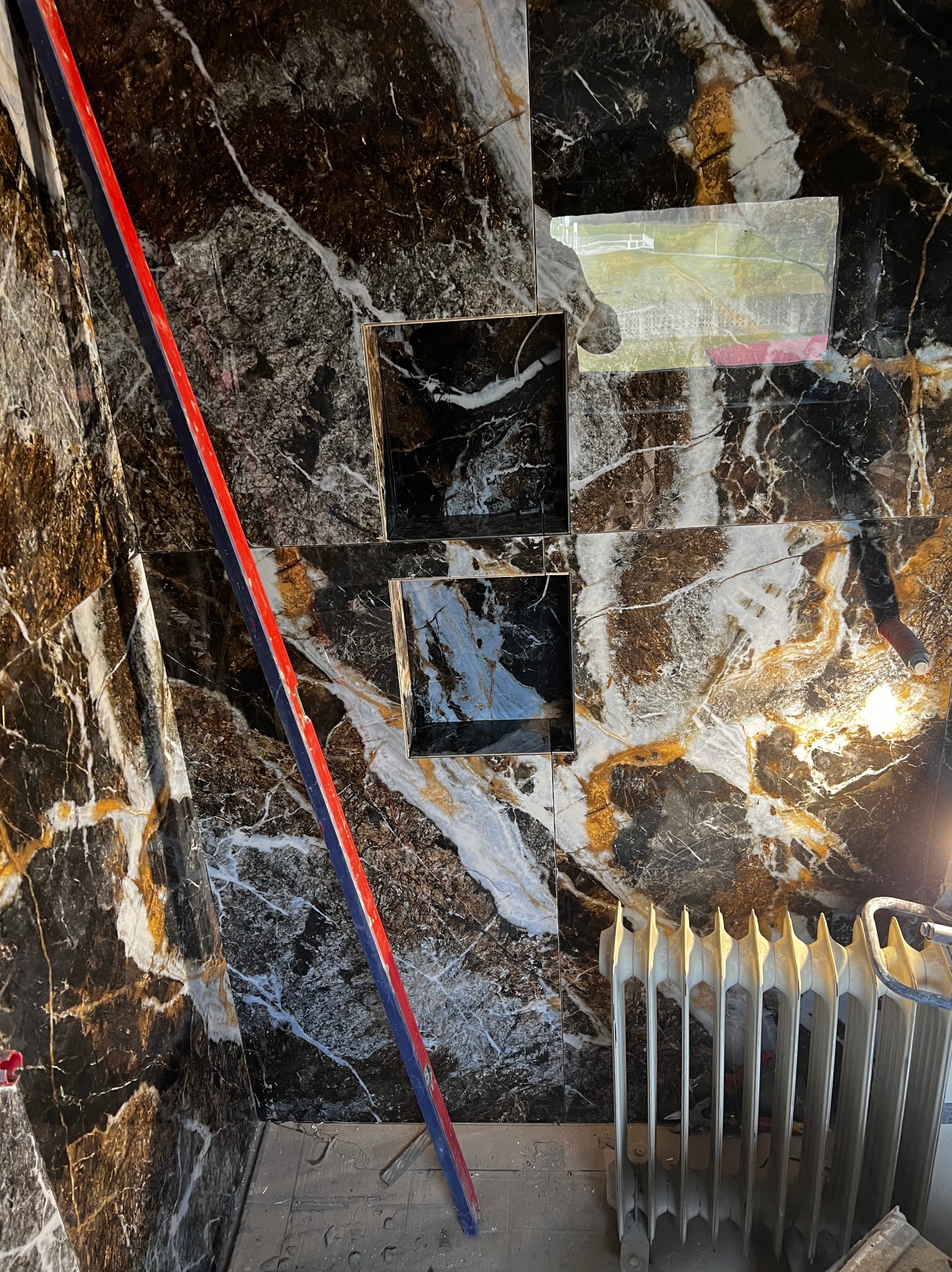 Ściana łazienki wyłożona czarno-białymi płytkami imitującymi marmur z złotymi żyłkami, z wnękami na kosmetyki, częściowo zaklejona folią ochronną, z widoczną poziomicą i grzejnikiem.