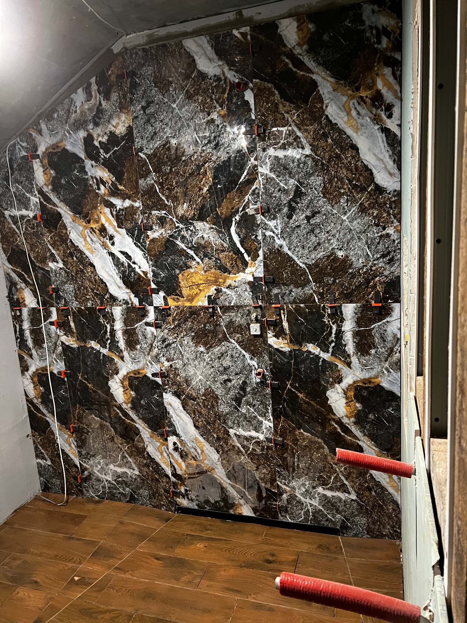 Ściana wyłożona płytkami imitującymi marmur w odcieniach czerni, bieli i złota, z widocznymi krzyżykami dystansowymi i poziomowaniem, podłoga wyłożona drewnopodobnymi płytkami, widoczne kable i rury.