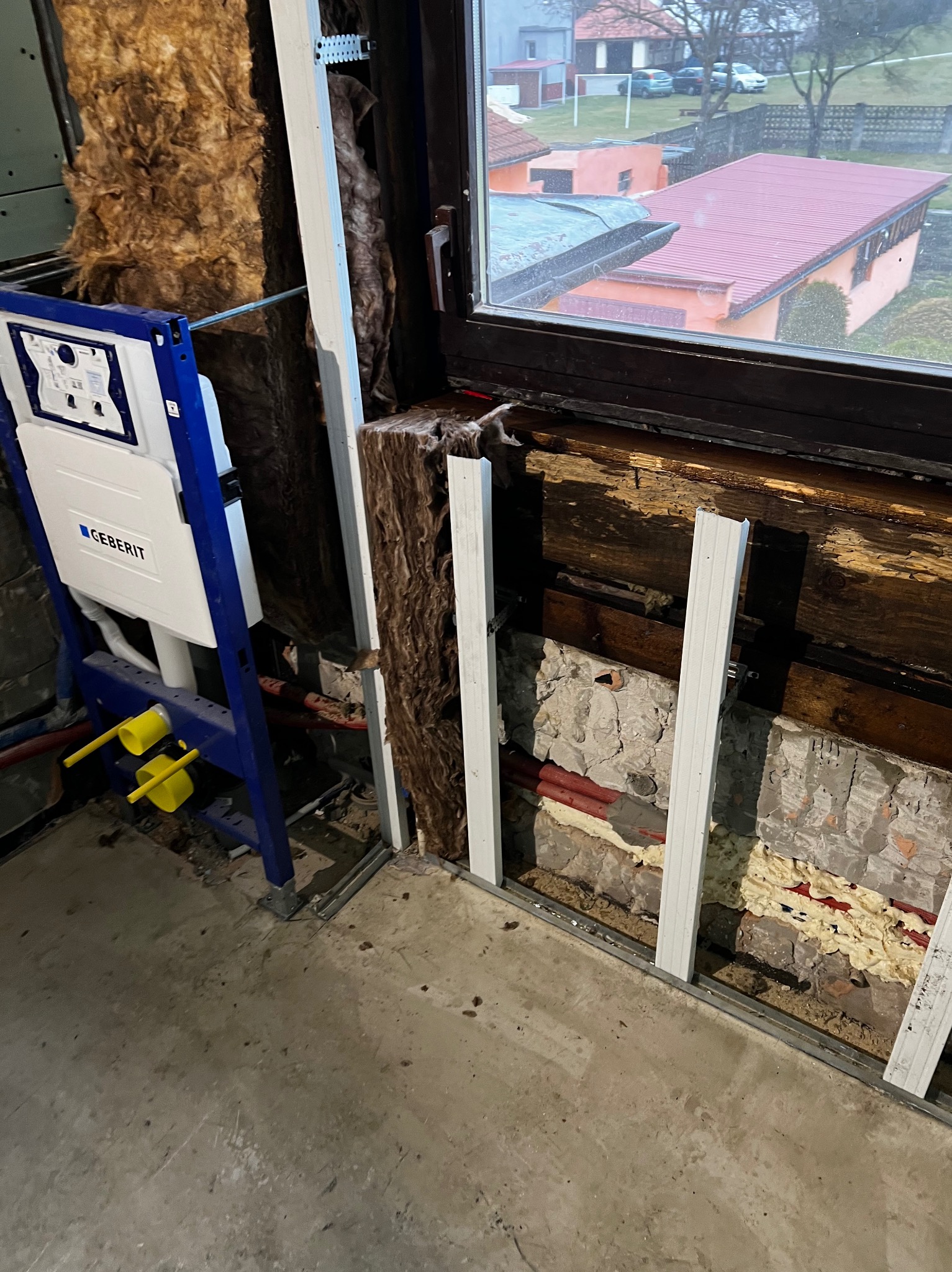 Stelaż podtynkowy Geberit zamontowany w łazience w trakcie prac wykończeniowych, widoczne ocieplenie ścian wełną mineralną i fragment okna z widokiem na dom z czerwonym dachem.