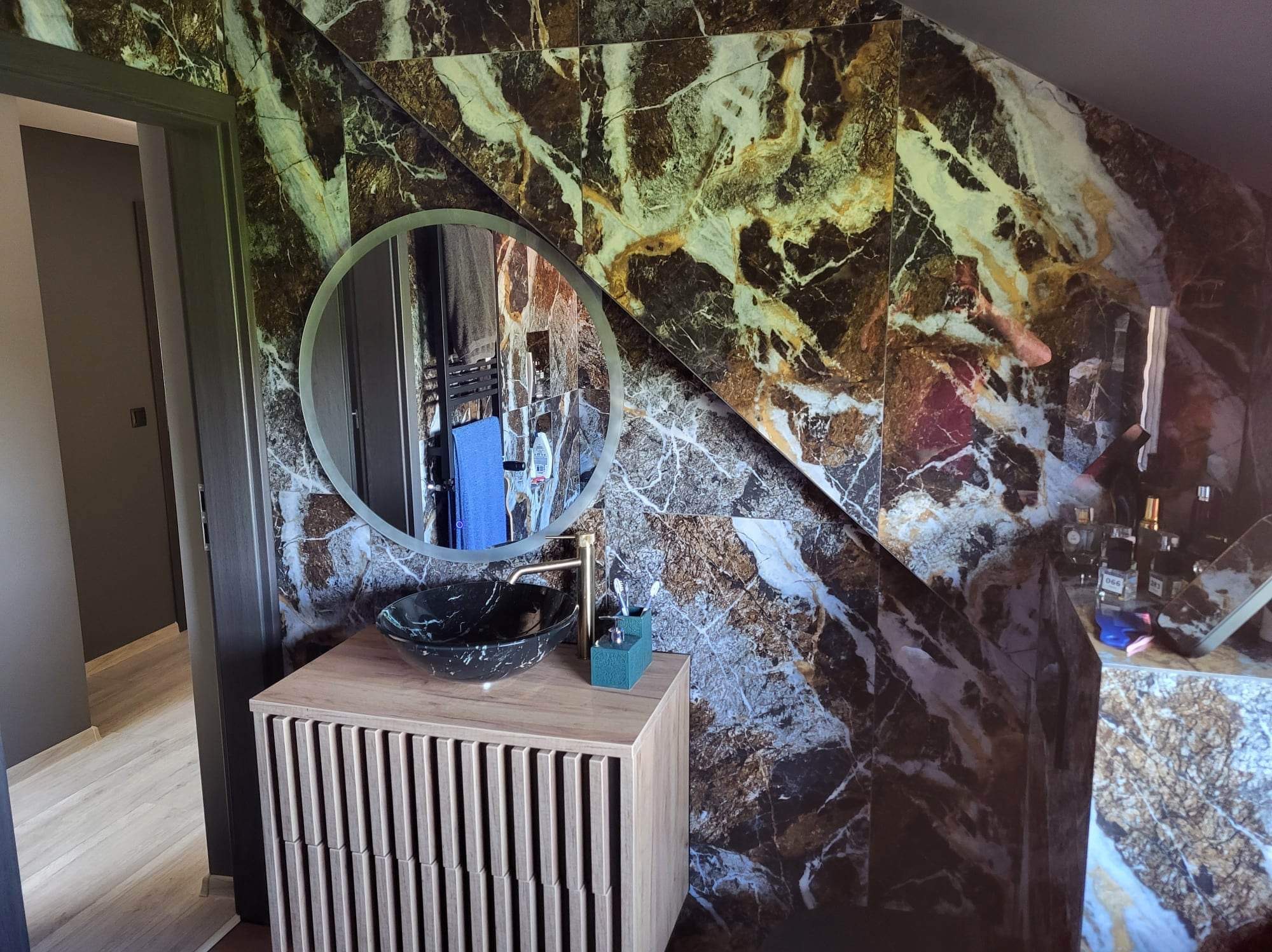 Nowoczesna łazienka z marmurowymi płytkami na ścianach, okrągłym lustrem i umywalką nablatową w kolorze czarnym z białymi żyłkami, szafka z drewnianymi lamelami.