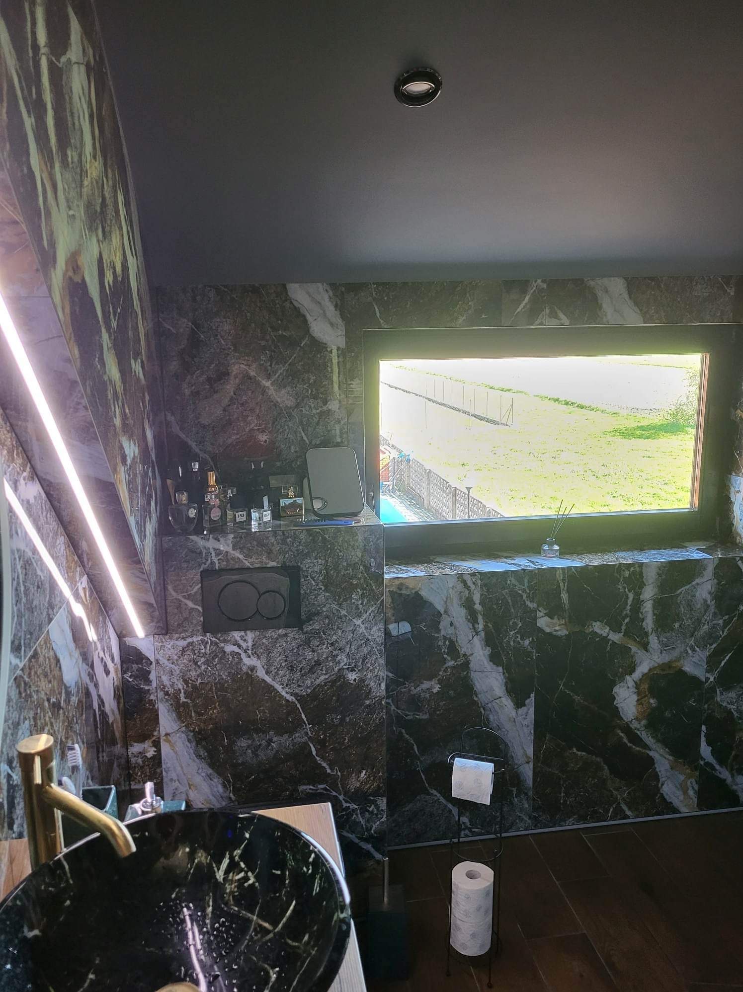 Elegancka łazienka w Opolu z czarnymi marmurowymi płytkami, złotą armaturą przy czarnej umywalce i widokiem na zieleń za oknem.