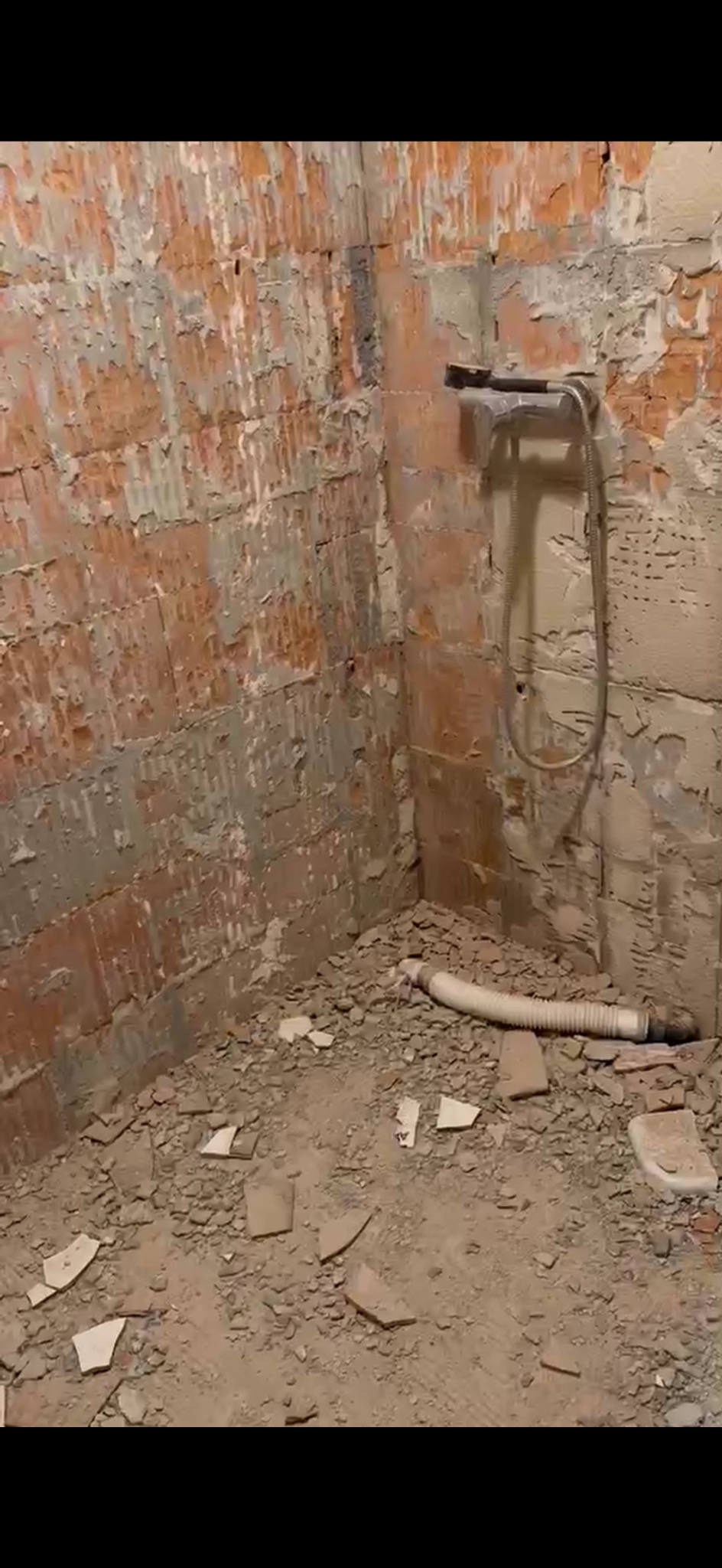 Łazienka w trakcie remontu: ściany z odsłoniętą cegłą, resztki starego tynku i gruzu na podłodze, zamontowana bateria prysznicowa i wąż odpływowy.