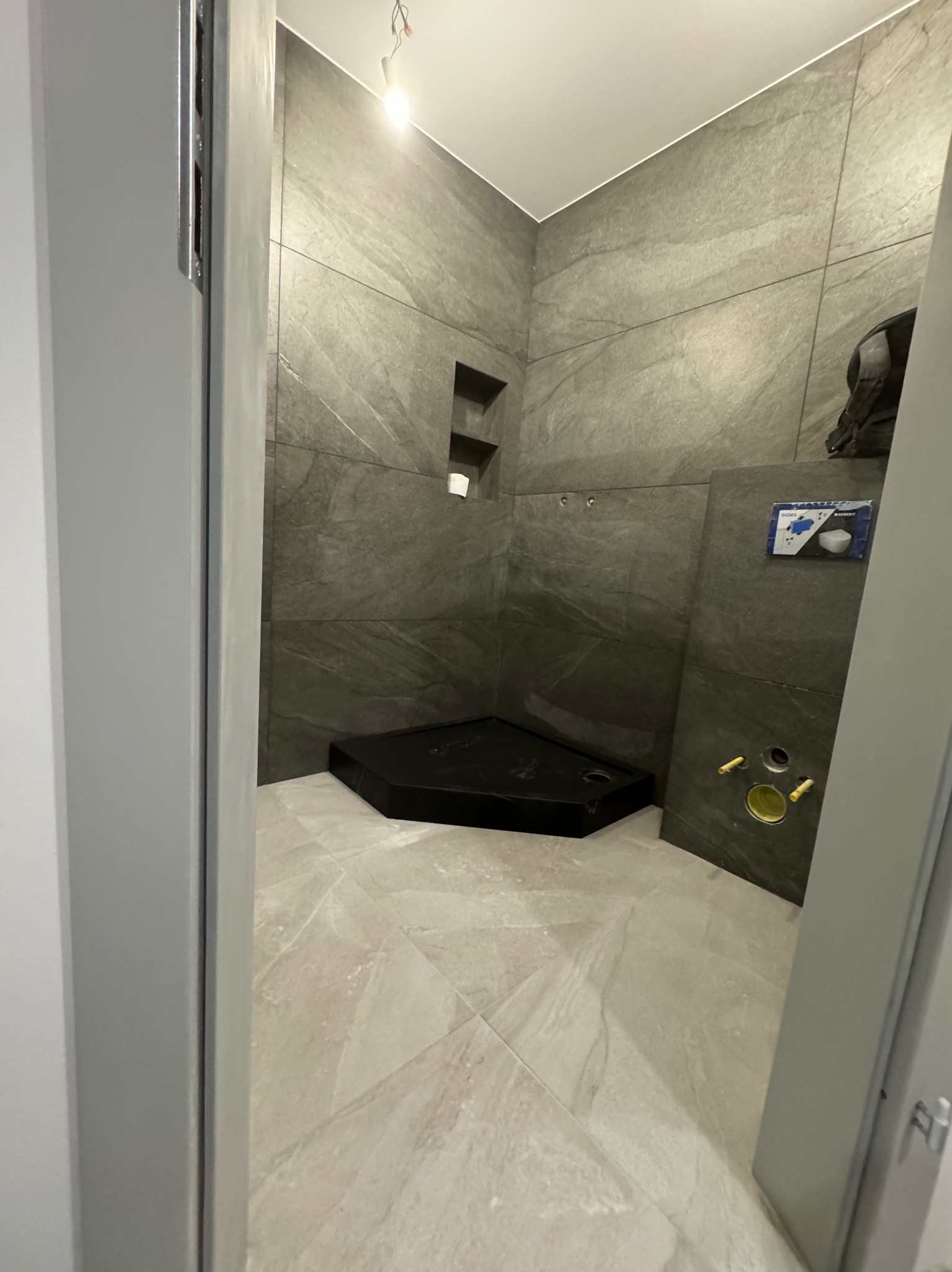 Częściowo wykończona kabina prysznicowa z ciemnoszarą okładziną ścienną imitującą kamień i jasnobeżową podłogą z płytek imitujących marmur, widoczny czarny brodzik i elementy instalacji podtynkowych.