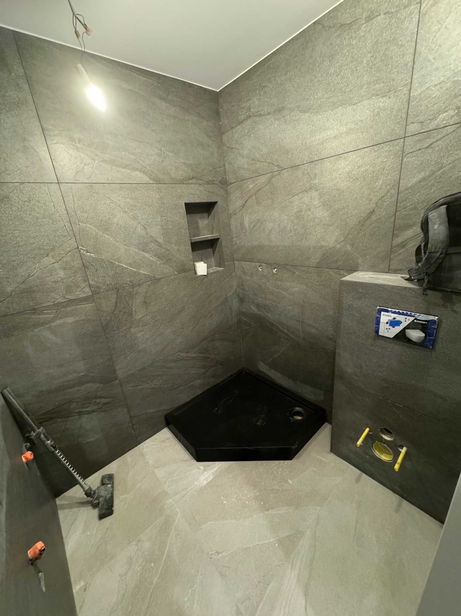 Wnętrze łazienki w trakcie prac wykończeniowych; widoczny czarny brodzik, szare płytki imitujące kamień na ścianach i podłodze, instalacja podtynkowego WC z przyciskiem spłukującym, oraz otwory...