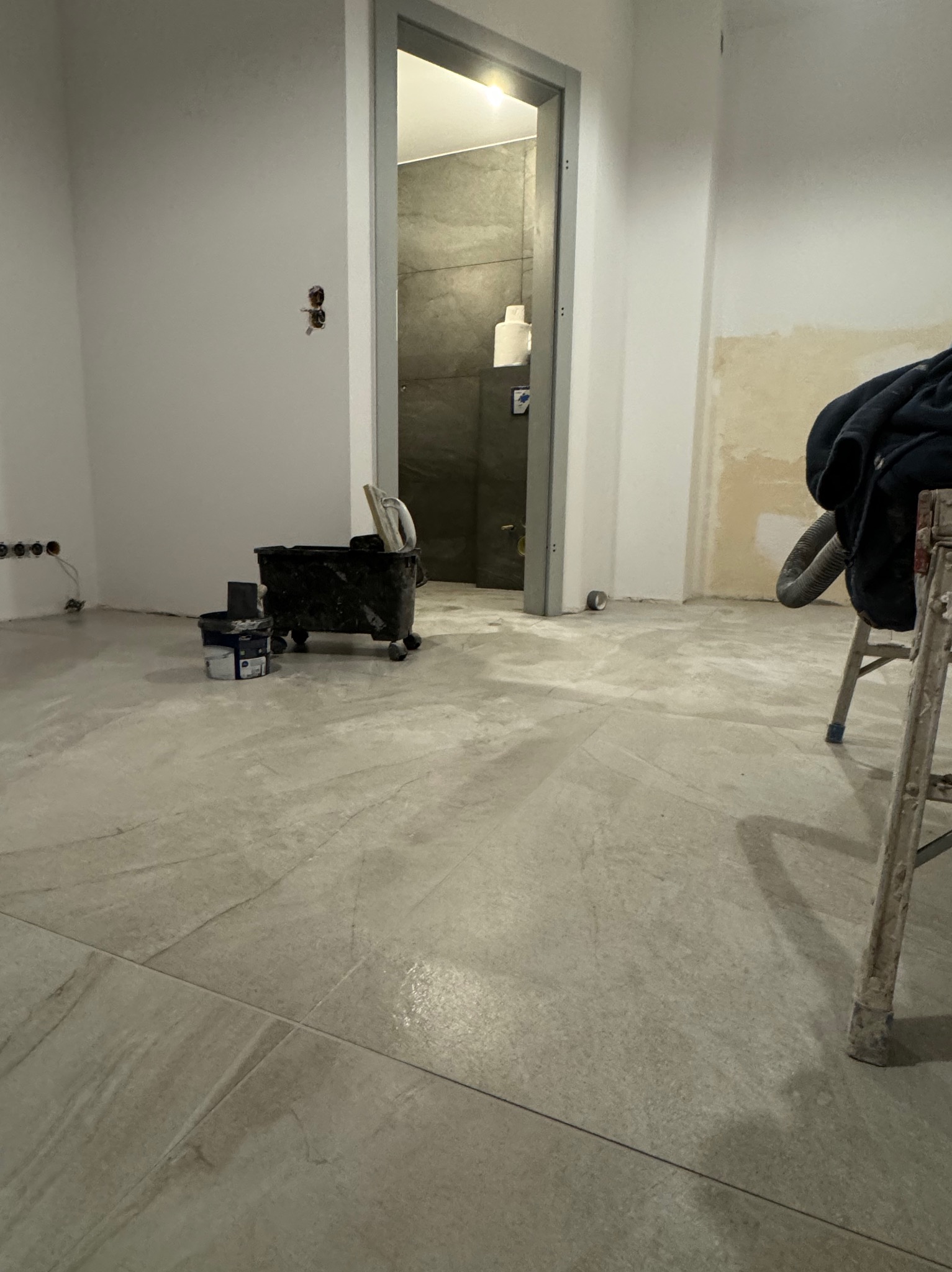 Przygotowania do prac wykończeniowych w Opolu: widok na podłogę z szarych płytek, wiadro z narzędziami malarskimi i drabina w tle łazienki z szarą okładziną.