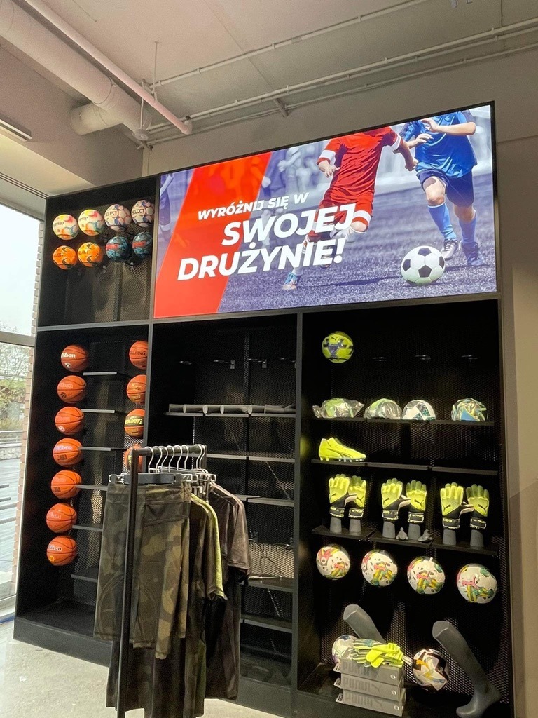 Ekspozycja w sklepie sportowym: piłki do piłki nożnej i koszykówki, rękawice bramkarskie, buty, odzież sportowa, a w tle ekran z hasłem 'Wyróżnij się w swojej drużynie!'