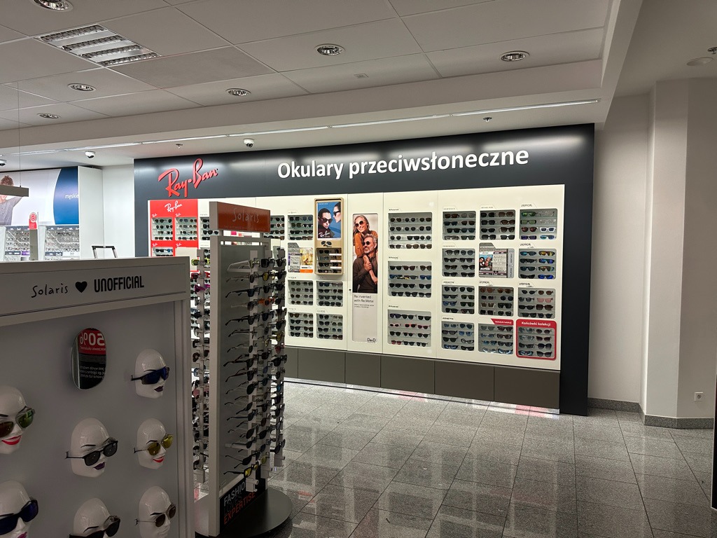 Ekspozycja okularów przeciwsłonecznych Ray-Ban w salonie optycznym, widoczne modele na stojakach i panelach ściennych.