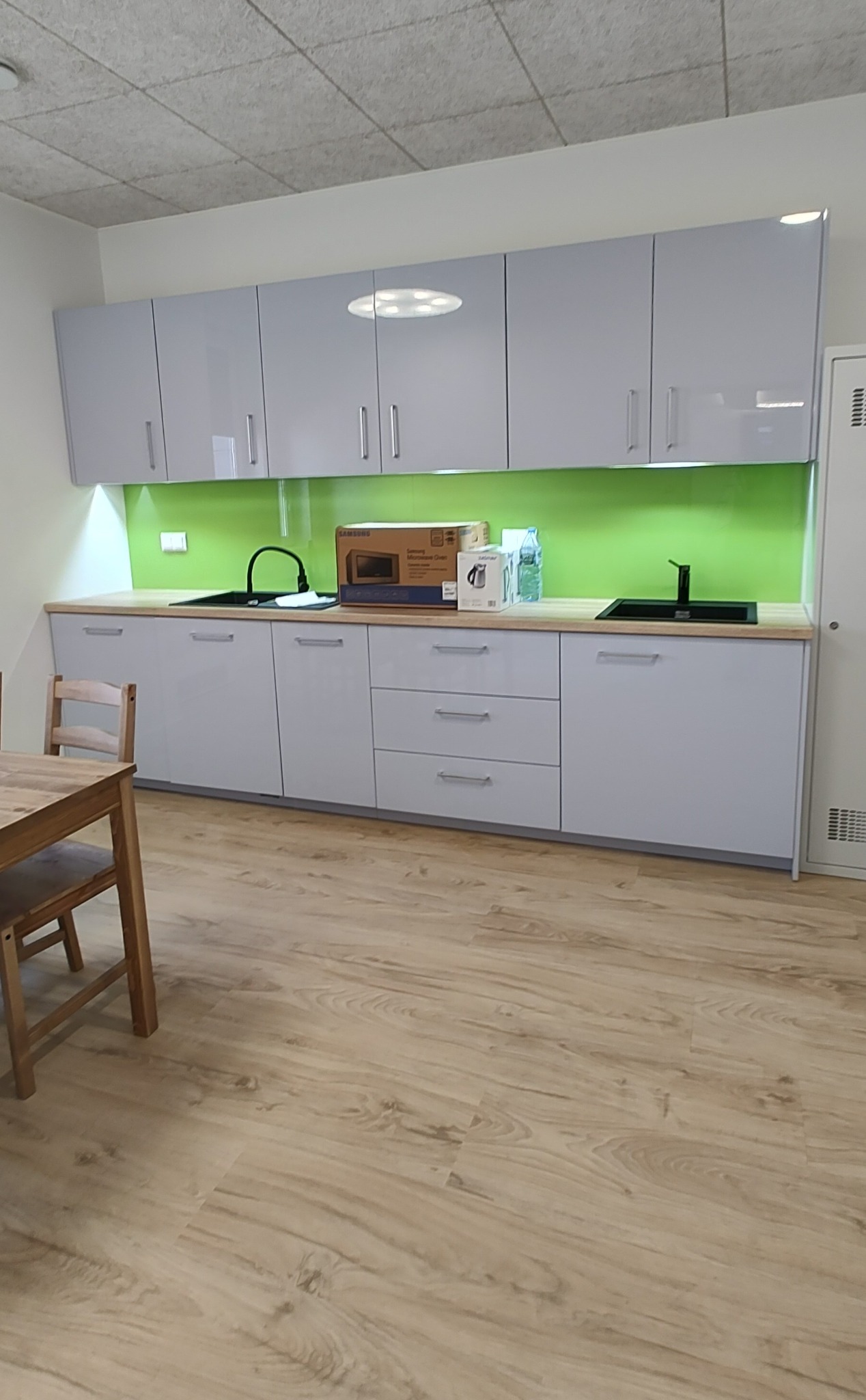 Jasnoszare szafki kuchenne z zielonym panelem, czarnym zlewem i armaturą, oraz drewnianym blatem, z pudełkami na blacie i stołem z krzesłami z przodu.