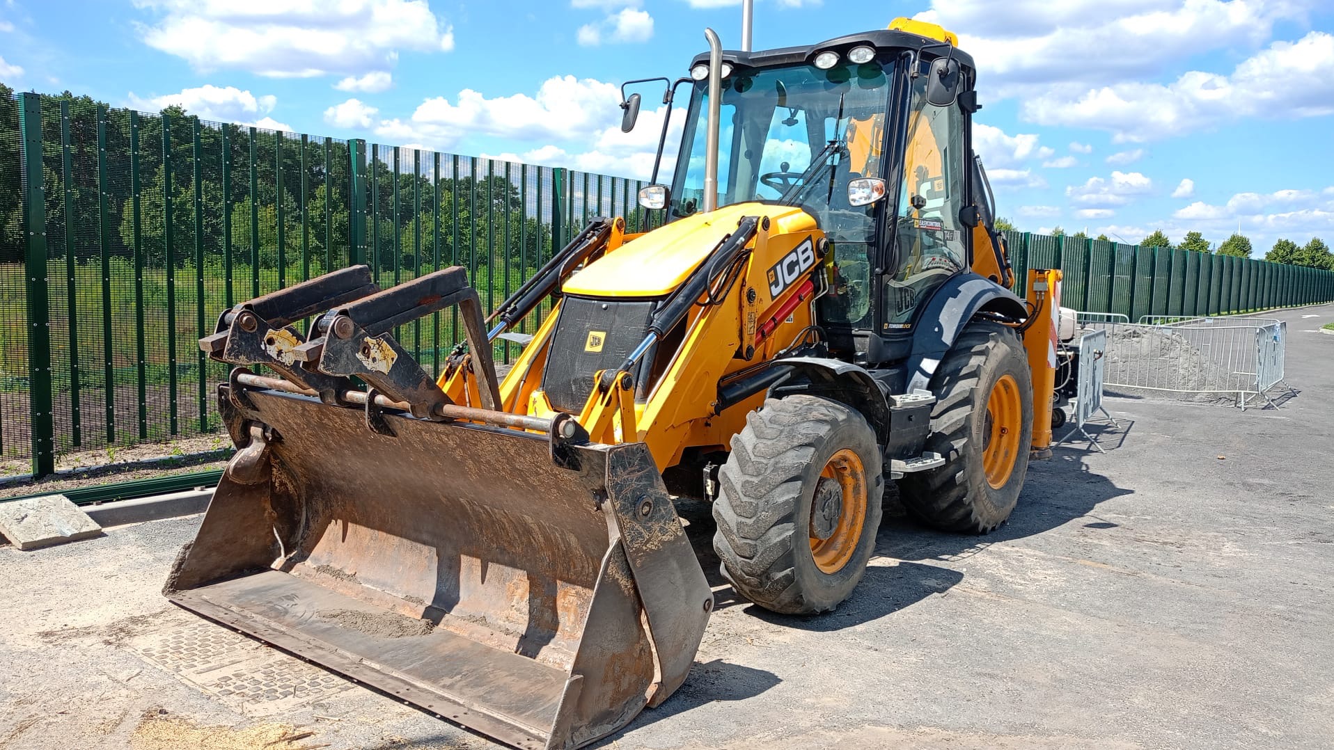 Żółto-czarna koparko-ładowarka JCB z wyraźnymi śladami użytkowania na łyżce, zaparkowana na asfaltowej powierzchni z zielonym ogrodzeniem w tle.