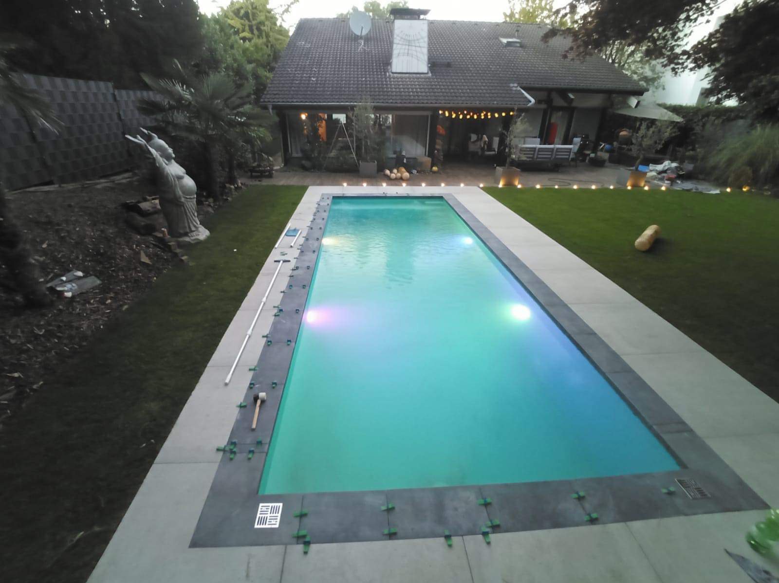 Narożny widok na basen z podświetleniem w trakcie wykończenia, widoczne narzędzia i elementy montażowe, w tle dom z ogrodem i dekoracjami.