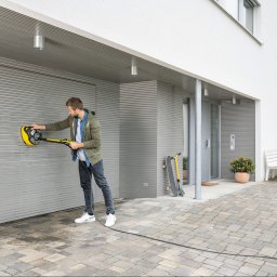 LFant Robert Mirowski - Mężczyzna w jeansach i zielonej bluzie czyści szarą, metalową bramę garażową za pomocą żółtej, okrągłej myjki ciśnieniowej Kärcher; widoczny fragment kostki brukowej, drzwi wejściowe i hulajnoga...