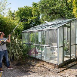 LFant Robert Mirowski - Mężczyzna w dżinsach i szarej koszulce myje szklarnię ciśnieniową myjką Kärcher z teleskopową lancą, otoczony bujną zielenią ogrodu.