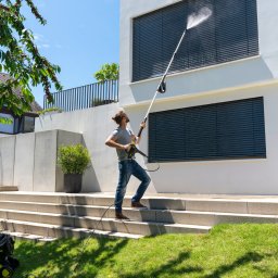 LFant Robert Mirowski - Mężczyzna w okularach, stojący na schodach, czyści okno na parterze nowoczesnego domu za pomocą myjki ciśnieniowej Kärcher. W tle trawnik i drzewo.