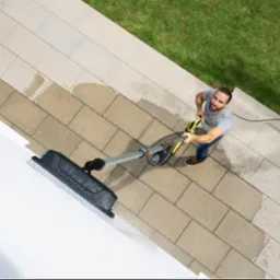 Mężczyzna z brodą, widziany z góry, czyści białą ścianę zewnętrzną za pomocą myjki ciśnieniowej z nasadką szczotkową, stojąc na kamiennej kostce brukowej obok trawnika.