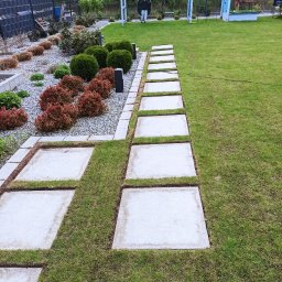 LFant Robert Mirowski - Ogród z trawnikiem, ścieżką z betonowych płyt i ozdobnymi rabatami z krzewami; w tle plac zabaw z huśtawką.