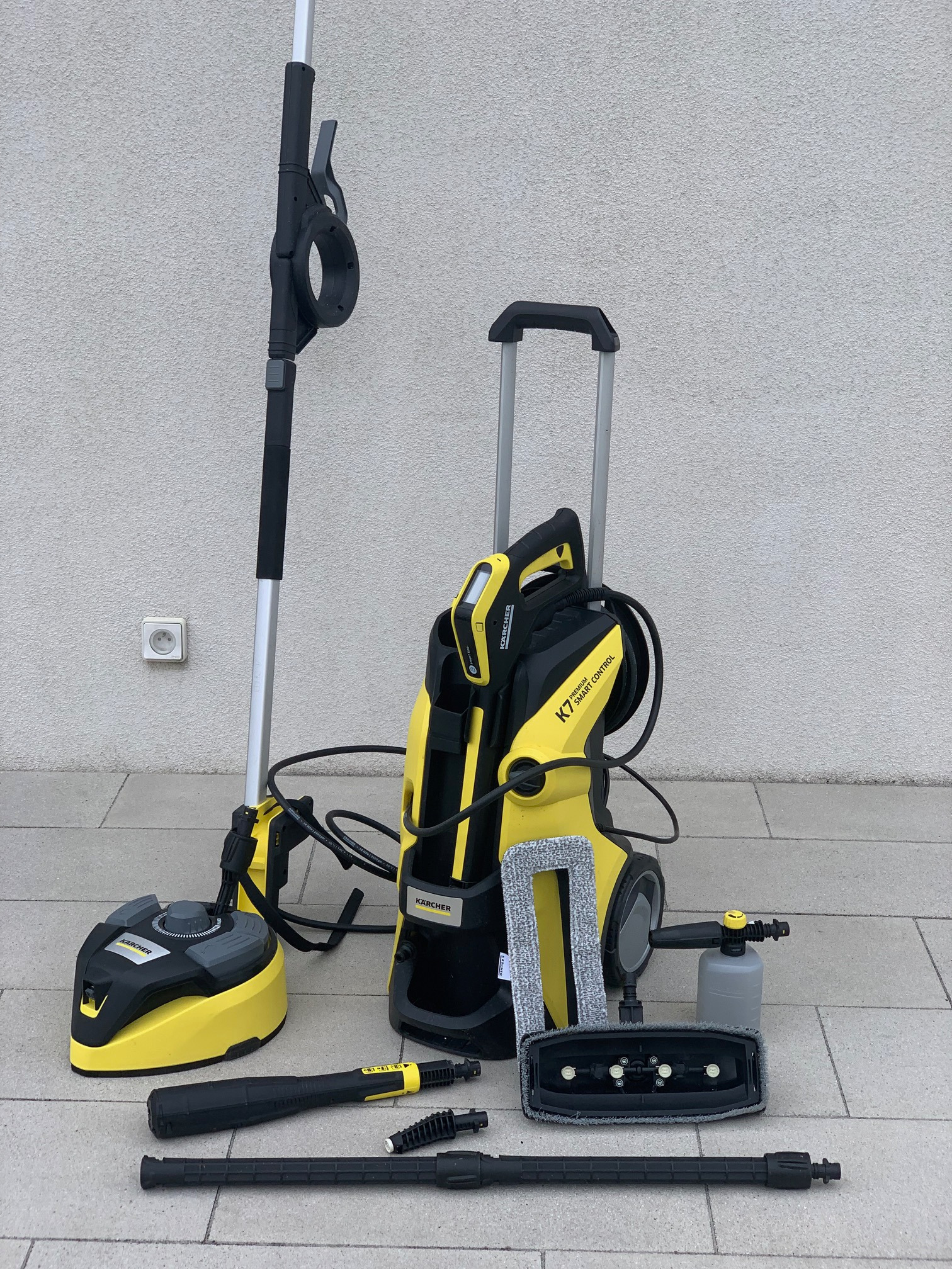 Zestaw do czyszczenia Kärcher K7 Smart Control z akcesoriami, w tym szczotka rotacyjna i przystawka do mycia okien, ustawiony na szarym bruku na tle jasnej ściany.