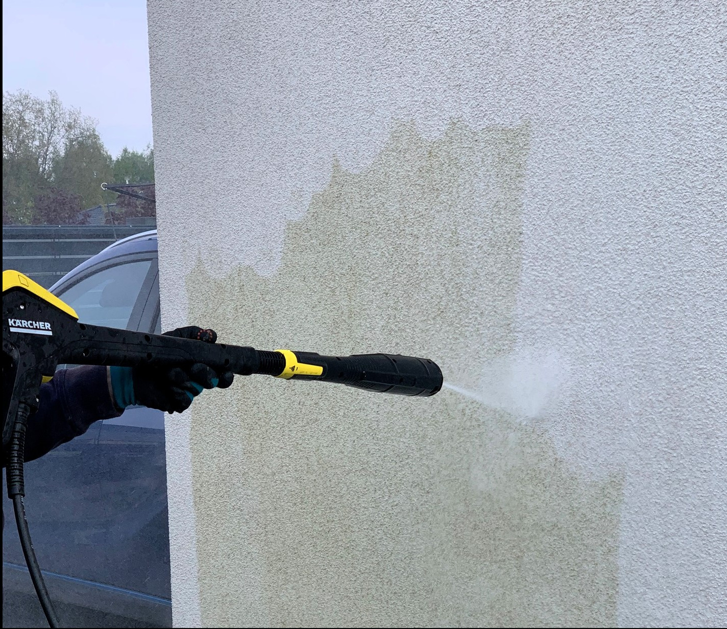 Czyszczenie elewacji budynku myjką ciśnieniową Kärcher, widoczne różnice w kolorze przed i po czyszczeniu, fragment samochodu w tle.