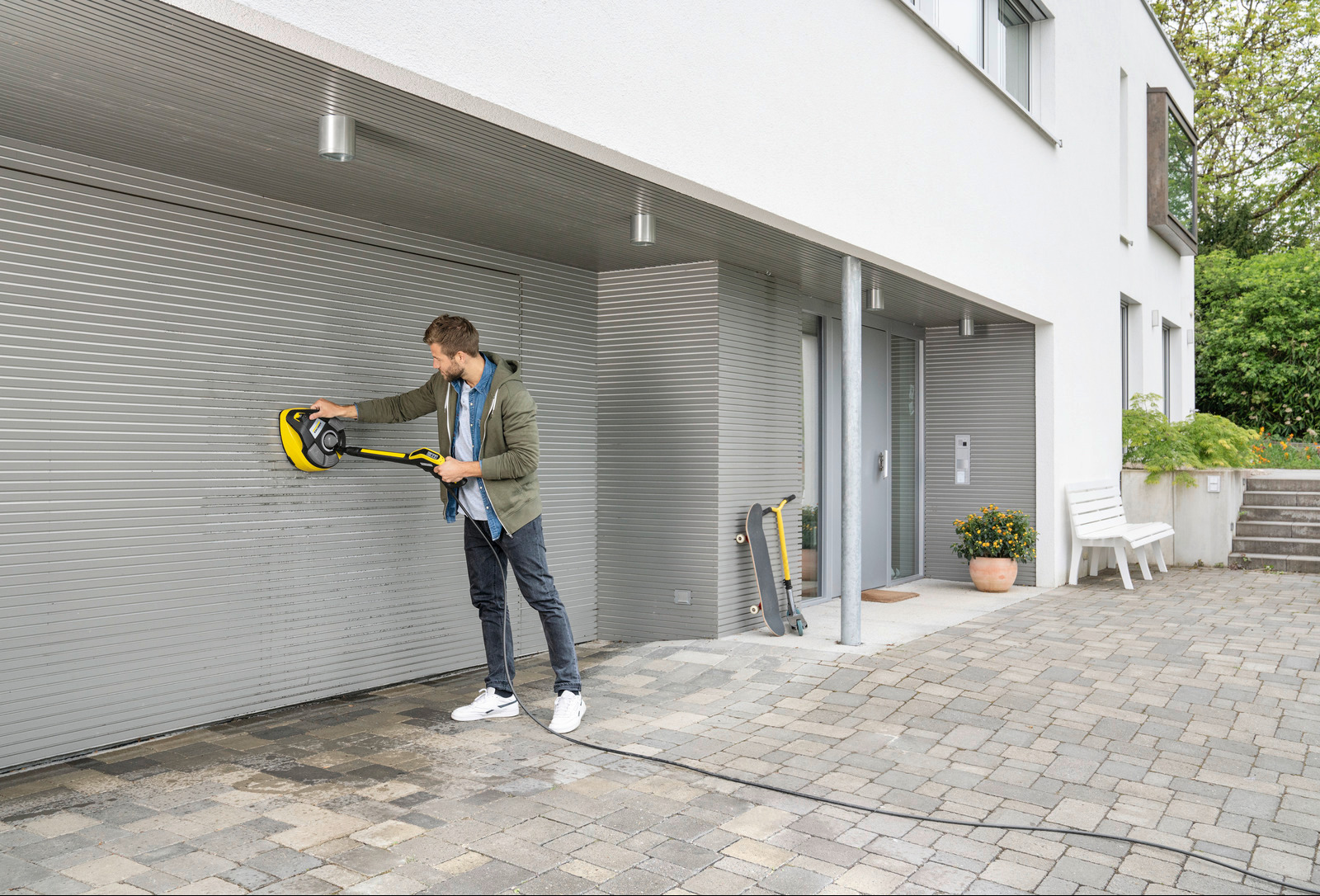 Mężczyzna w jeansach i zielonej bluzie czyści szarą, metalową bramę garażową za pomocą żółtej, okrągłej myjki ciśnieniowej Kärcher; widoczny fragment kostki brukowej, drzwi wejściowe i hulajnoga...