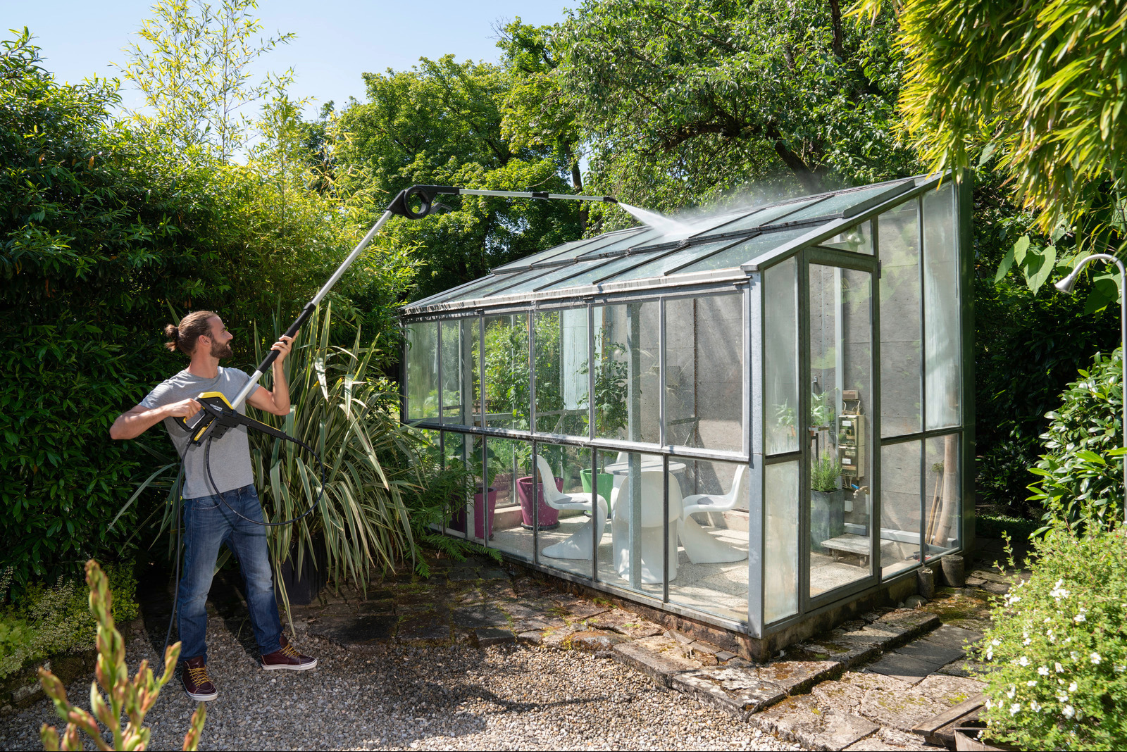 Mężczyzna w dżinsach i szarej koszulce myje szklarnię ciśnieniową myjką Kärcher z teleskopową lancą, otoczony bujną zielenią ogrodu.