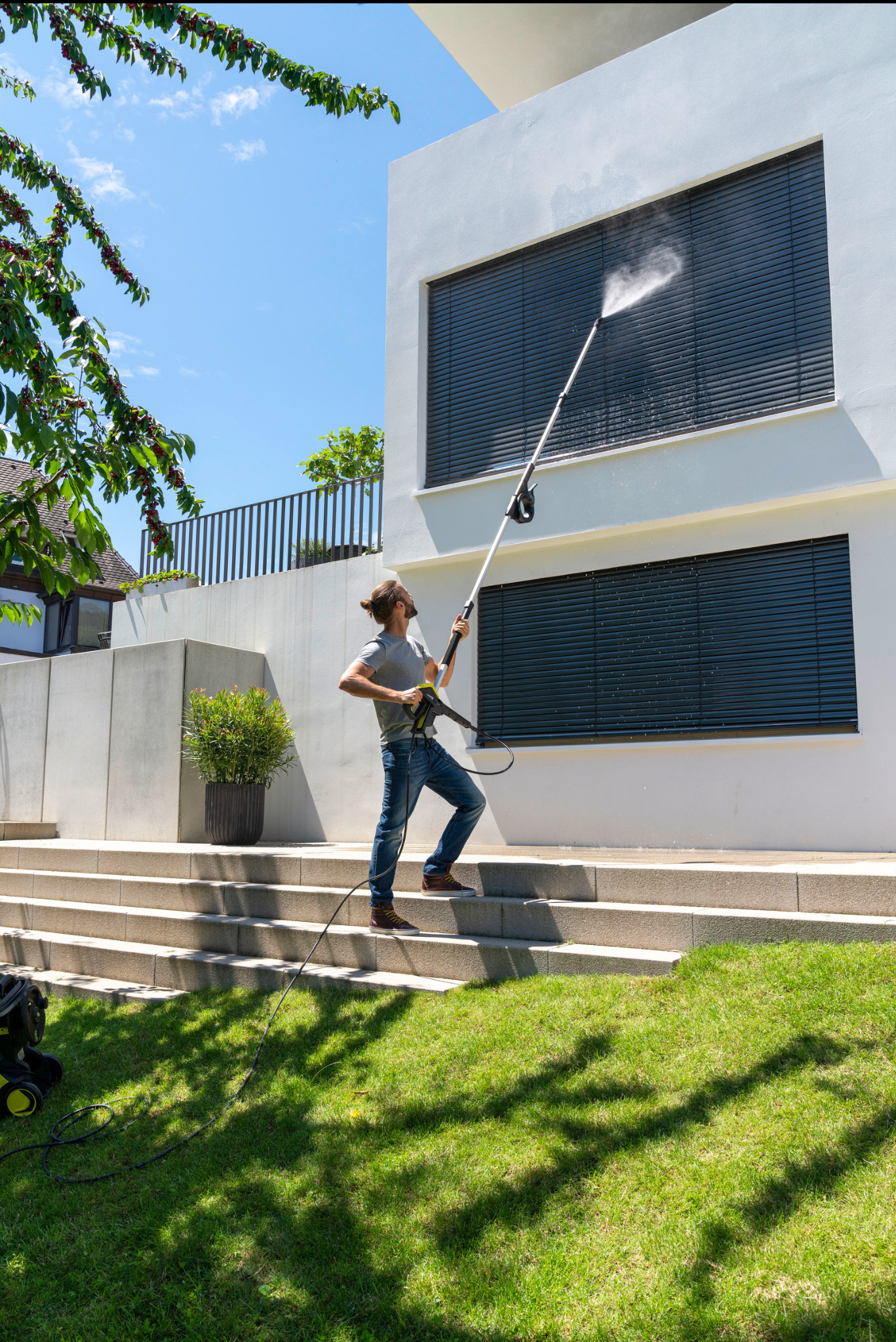 Mężczyzna w okularach, stojący na schodach, czyści okno na parterze nowoczesnego domu za pomocą myjki ciśnieniowej Kärcher. W tle trawnik i drzewo.