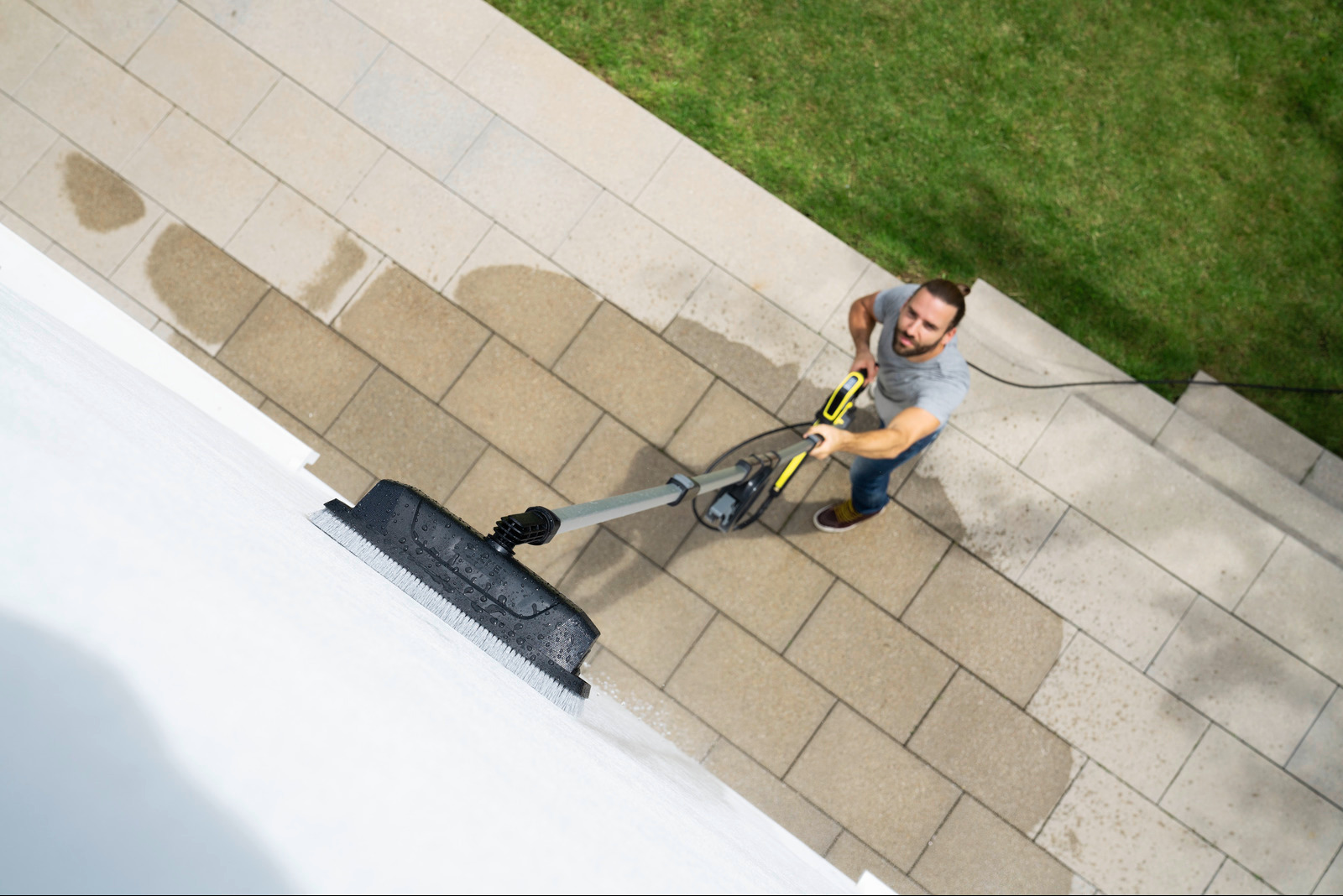 Mężczyzna z brodą, widziany z góry, czyści białą ścianę zewnętrzną za pomocą myjki ciśnieniowej z nasadką szczotkową, stojąc na kamiennej kostce brukowej obok trawnika.