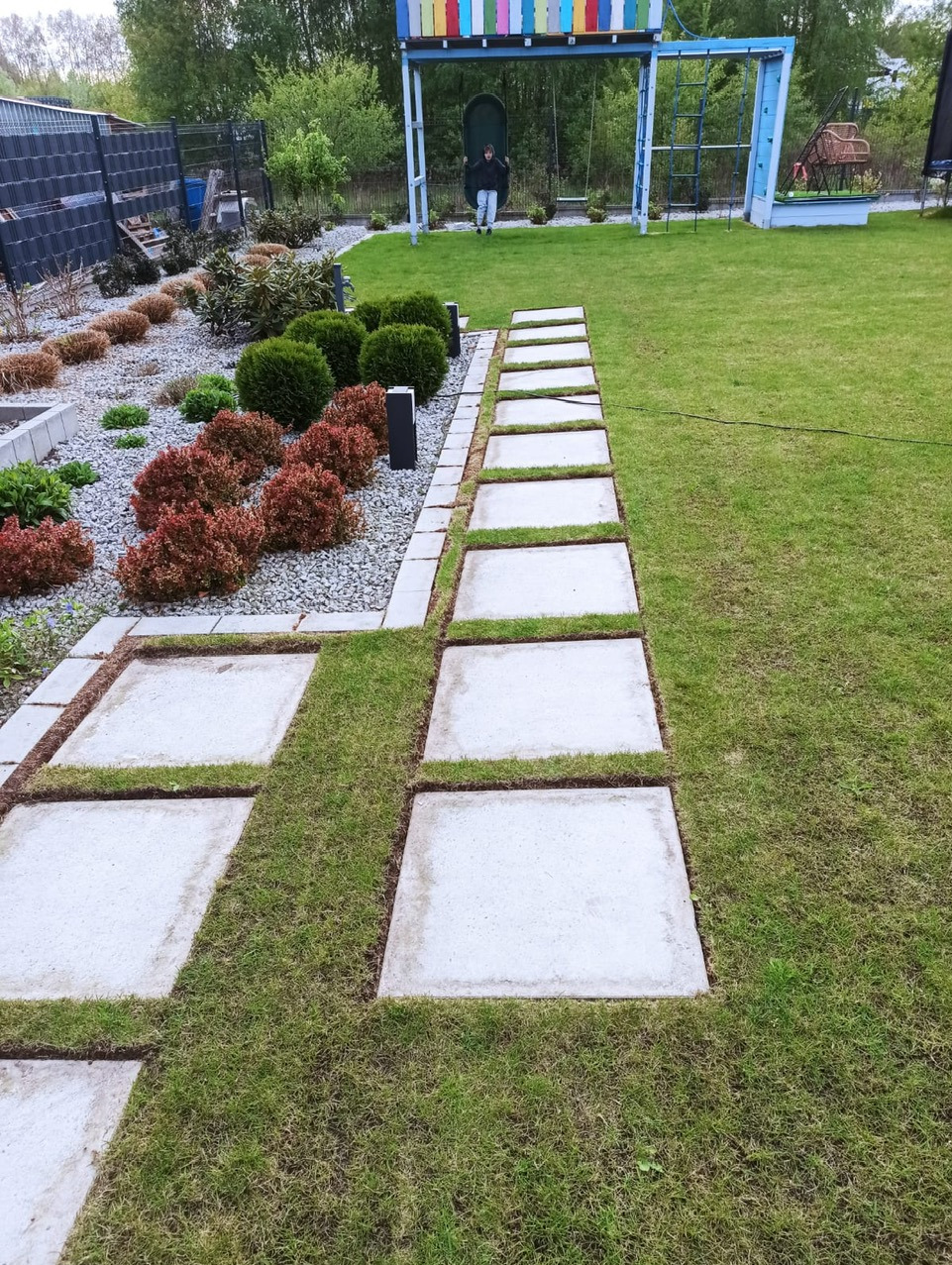 Ogród z trawnikiem, ścieżką z betonowych płyt i ozdobnymi rabatami z krzewami; w tle plac zabaw z huśtawką.