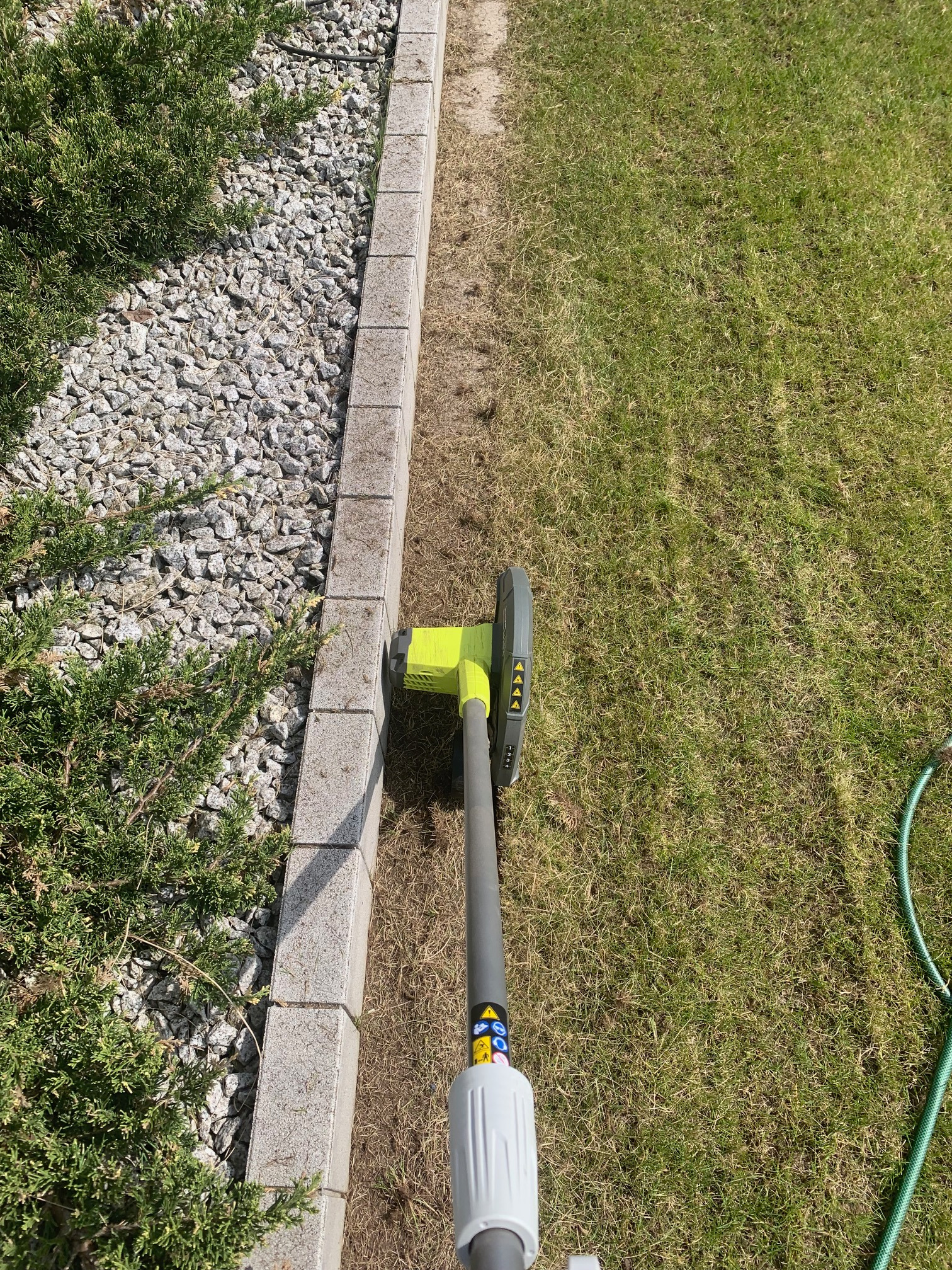 Elektryczny podkaszacz do trawy z żółtą głowicą przycinający trawę przy betonowym obrzeżu rabaty z krzewami i szarym żwirem, widok z góry.