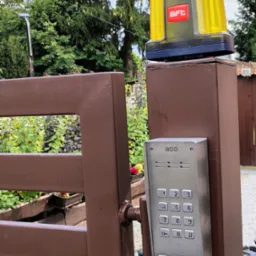 Brązowa brama z automatycznym systemem dostępu ACO, lampą ostrzegawczą BFT i widocznym ogrodem w tle.