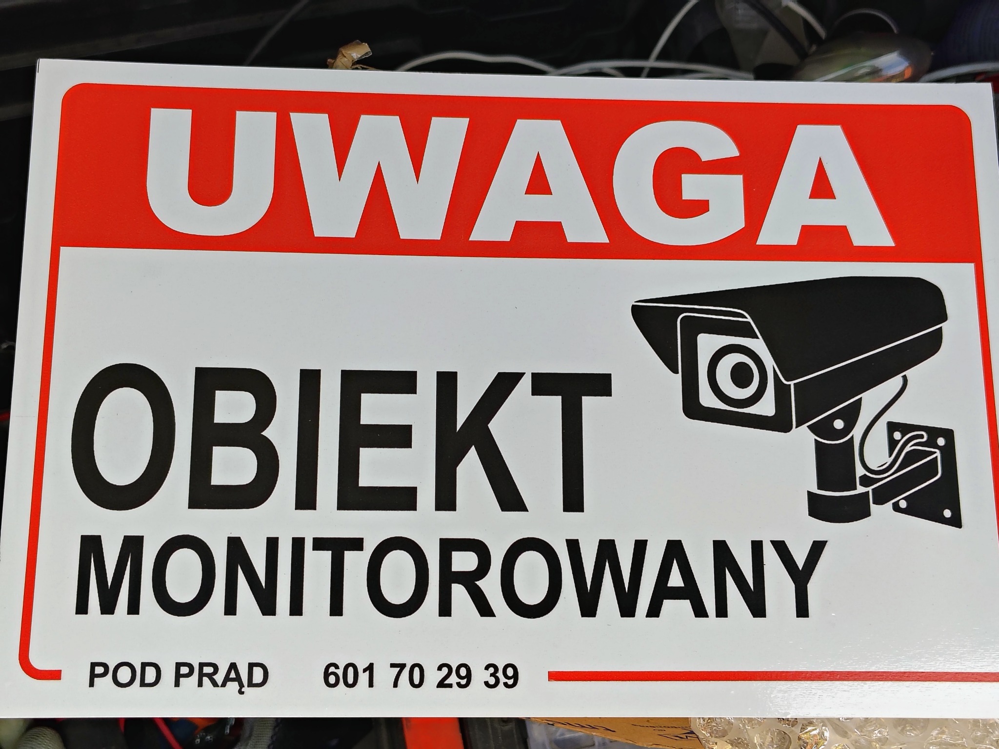 Tablica ostrzegawcza z napisem 'UWAGA OBIEKT MONITOROWANY' i grafiką kamery przemysłowej, pod spodem numer telefonu.