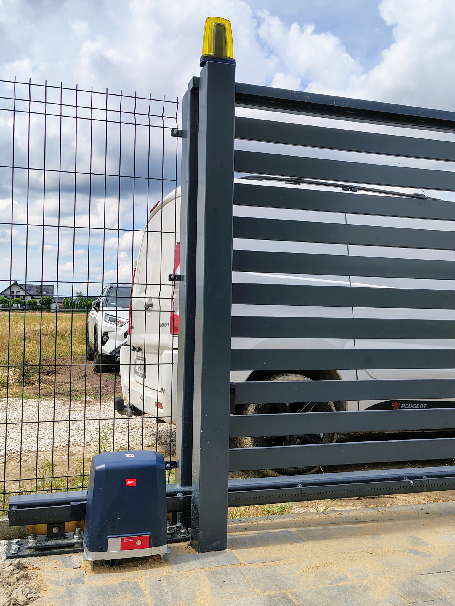 Nowoczesna, przesuwna brama z poziomych paneli w kolorze grafitowym, z widocznym napędem BFT Ultra i lampą ostrzegawczą, zamontowana na posesji z siatką ogrodzeniową i zaparkowanymi samochodami.
